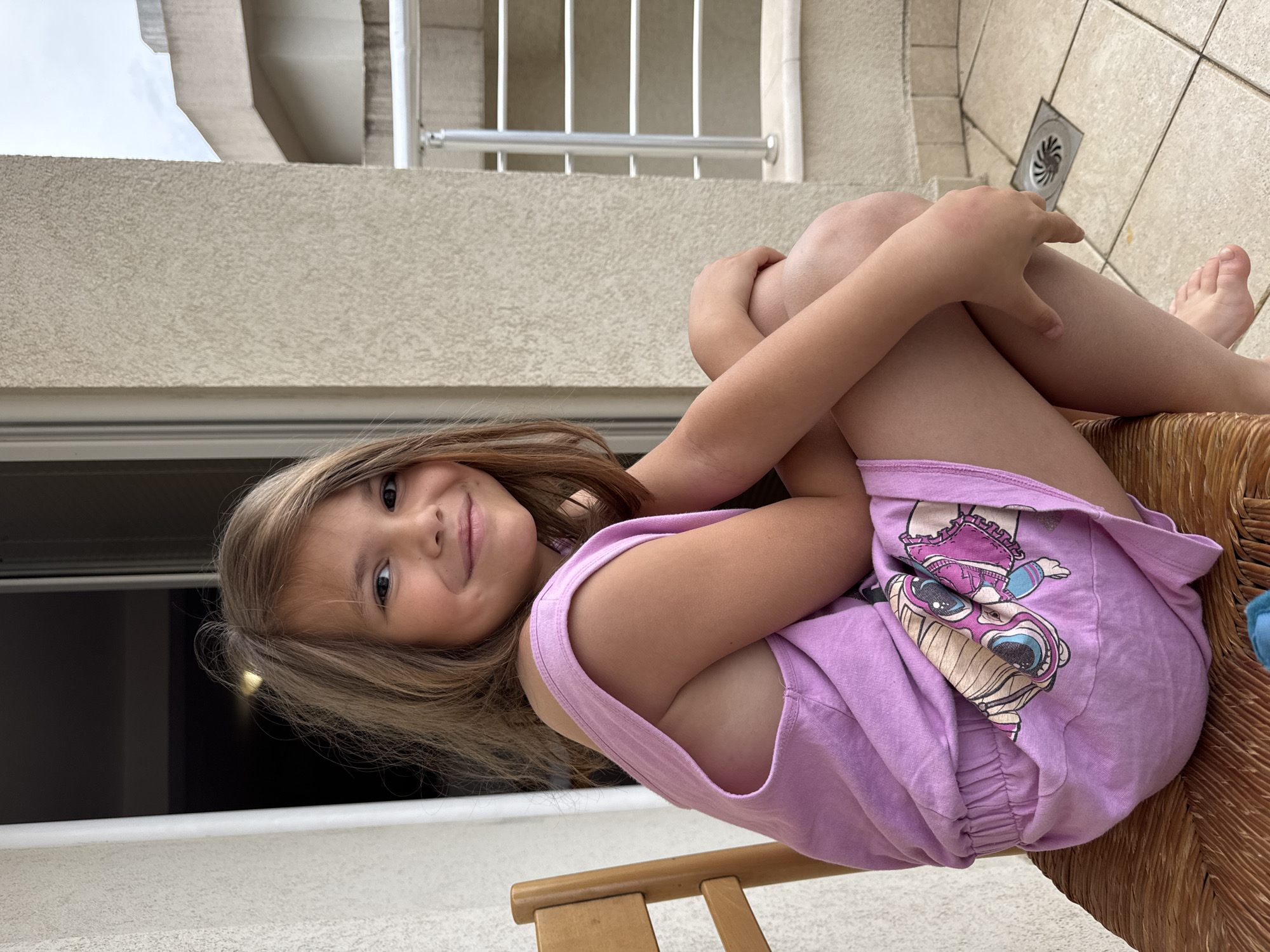 A young girl is lying on her side on a light brown wicker chair or lounge, smiling towards the camera. Her knees are drawn up towards her chest, and her arms are wrapped around her shins. She has long, light brown hair and a cheerful expression.
She is wearing a light purple tank top or casual dress that features a colorful graphic print on the front. The graphic depicts a stylized character, possibly resembling an LOL Surprise doll, with large pink and blue sunglasses and blonde pigtails.
The setting appears to be an outdoor balcony or terrace in Budva, Montenegro. To the left (which would be above if the image were rotated upright), there is a textured, light-colored concrete wall, above which parts of a building structure, a metal railing, and an overcast sky are visible. To the right (which would be below if rotated upright), a tiled floor with light-colored, possibly beige or cream, tiles is present, and a square metal drain is visible on the floor.
The lighting is soft, suggesting an overcast day during daylight hours. There is no discernible text visible in the image, other than the stylized character on her clothing. The overall mood of the image is happy and relaxed.