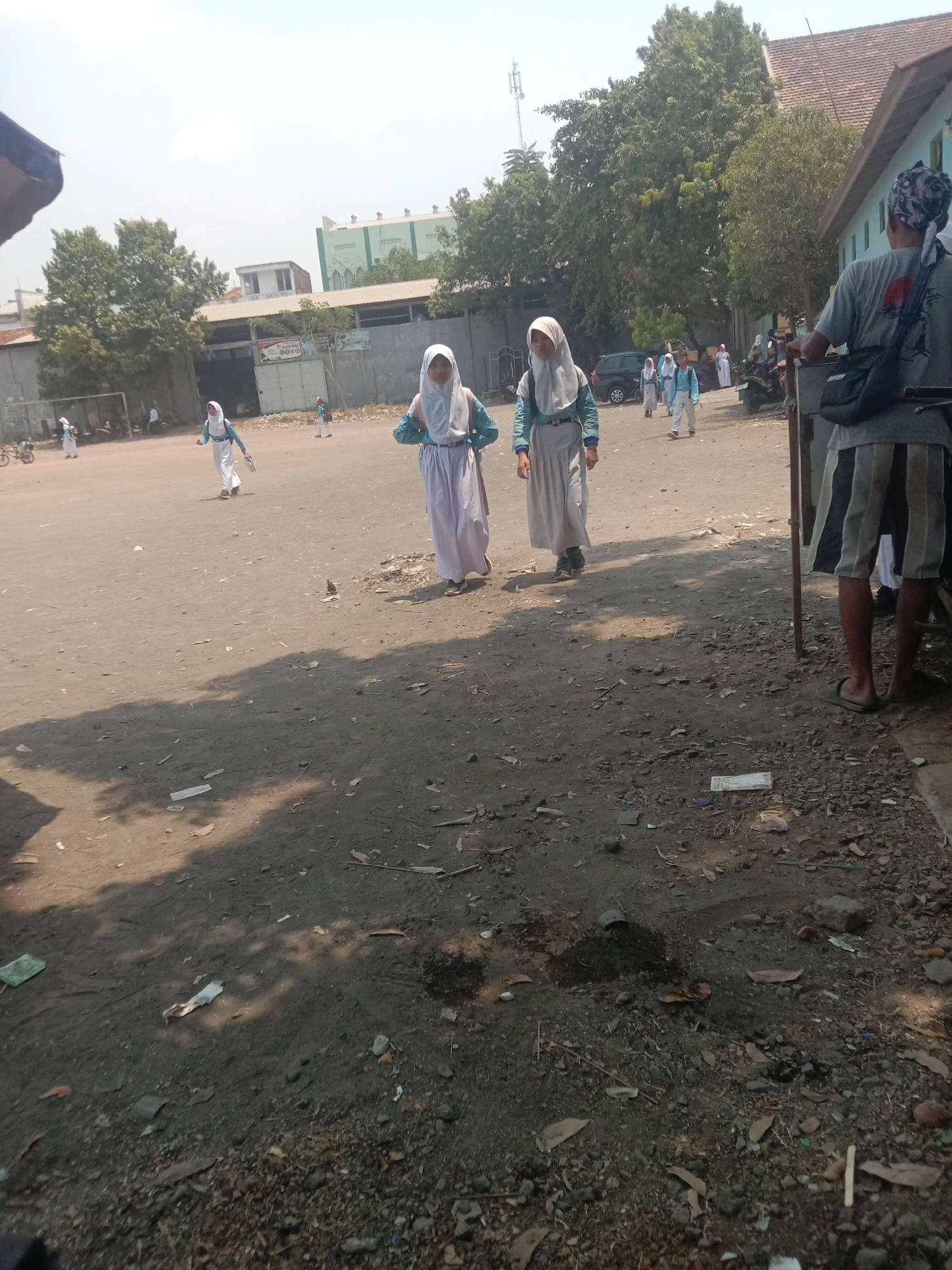 The image depicts an outdoor scene, likely in the city of Ngempit, Indonesia, with a predominantly dusty, light-brown ground surface. In the mid-ground, two young females wearing white hijabs and long, white skirts with light blue accents are walking from right to left, seemingly participating in school activities. They both carry books or papers. To the right of the frame, a person stands facing away from the camera, wearing a grey t-shirt with a red circular design on the back, dark shorts with vertical white stripes, and flip-flops. This individual is positioned near a wooden post. In the background, there are buildings, including one with a light green facade and Islamic architectural elements, as well as trees and a cell tower. The overall environment suggests a dry, possibly arid, area with scattered debris on the ground, including leaves, small stones, and some litter. The lighting indicates it is daytime, with shadows visible on the ground.