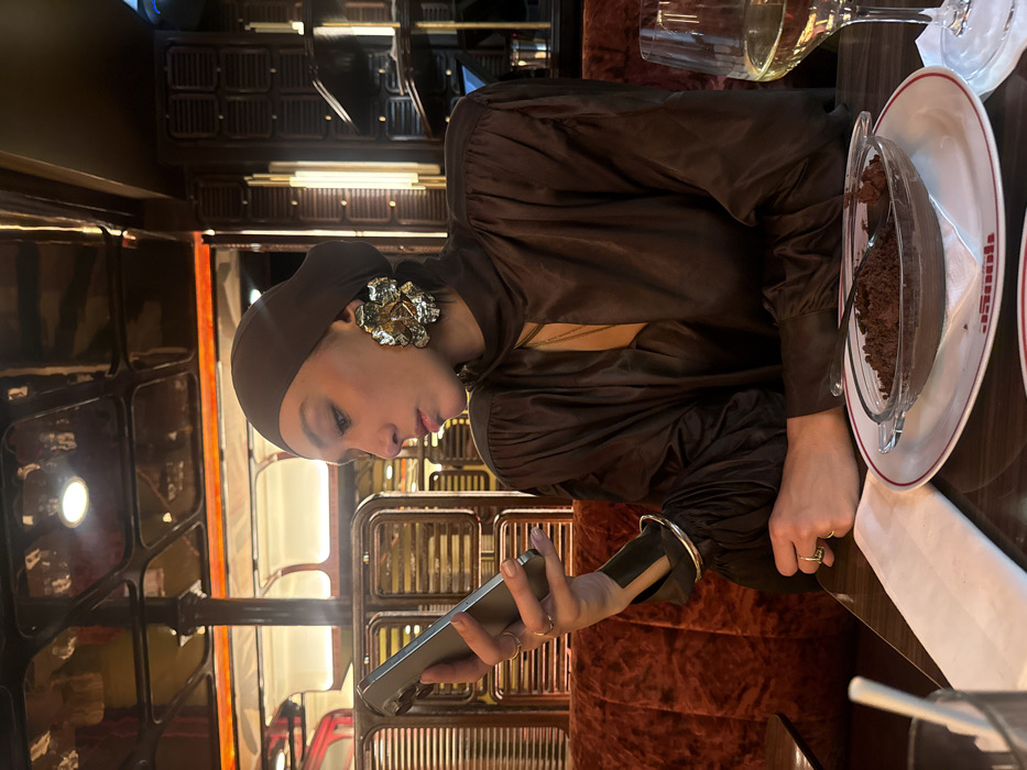 A woman in a dark brown headscarf and oversized, silver floral earrings holds a phone. She's wearing a brown, long-sleeved dress or top. A dessert dish with what looks like chocolate mousse or pudding is on the table to her right. A partially filled wine glass is also visible. The background features dark wood detailing and metal accents, creating a vintage or old-world ambiance.