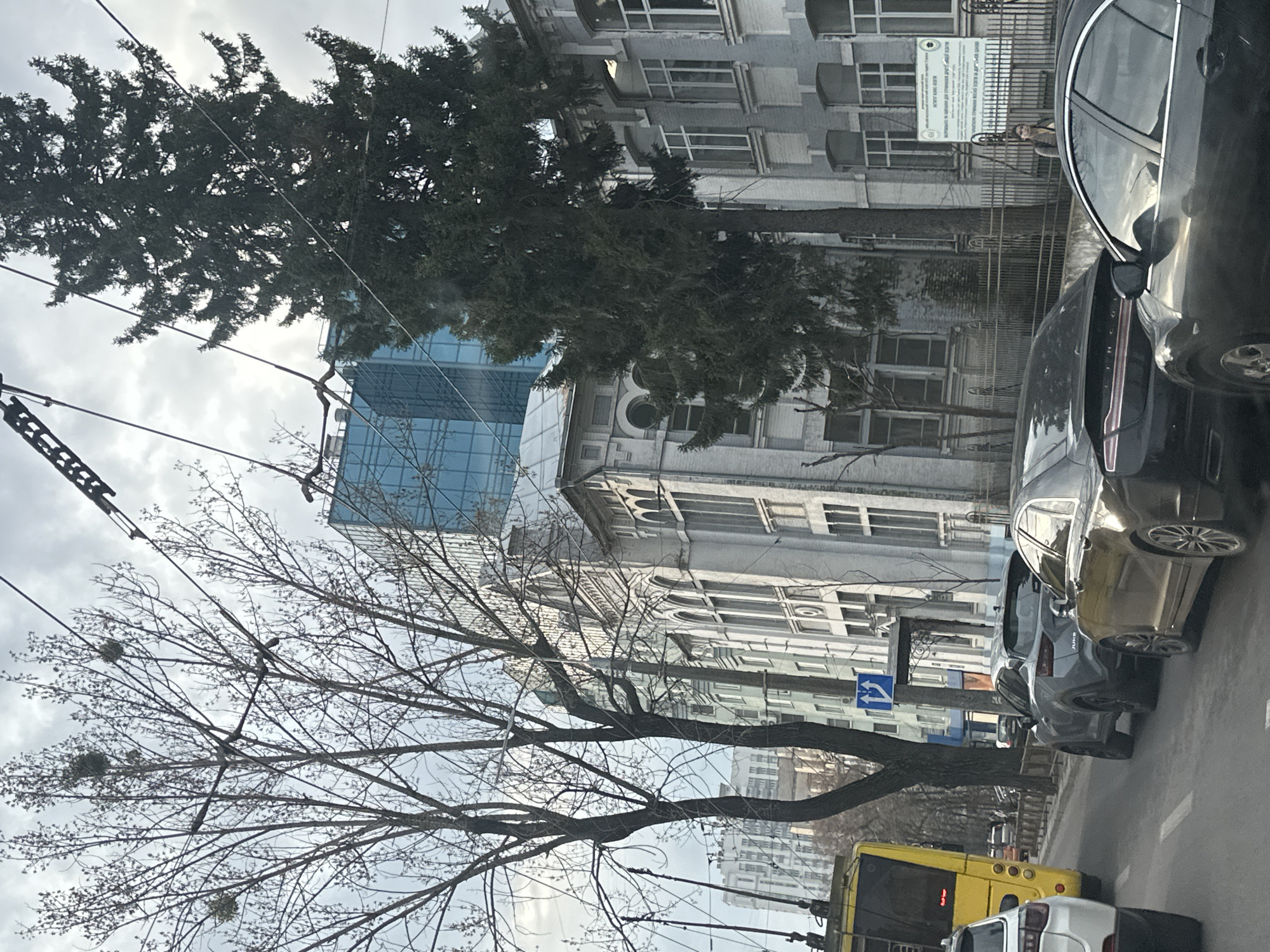 The image depicts a street scene in Kyiv, Ukraine. The view is likely from a vehicle. Cars are parked along the street. A yellow bus is visible. Tall buildings line the street, including older, light-colored buildings and a modern glass-faced structure. Bare trees and evergreens stand between the buildings and the street. Overhead power lines are present. A street sign indicates directions. A person stands behind a metal fence in the background.