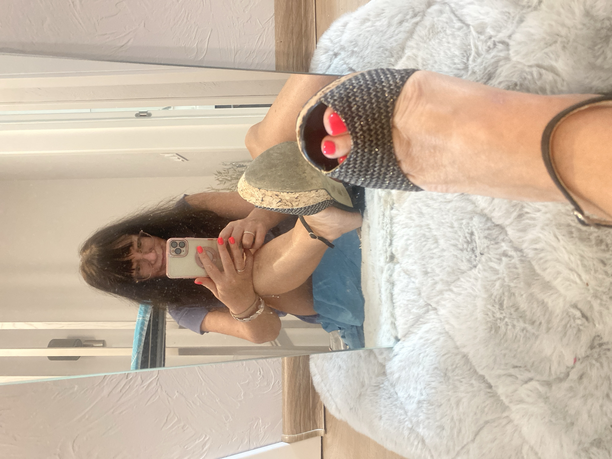 The media file is a selfie taken in a mirror, reflecting a woman. She has long, dark hair and is wearing glasses and a purple t-shirt. She is holding a smartphone, with which she is taking the photo, in her right hand. Her fingernails are painted a bright coral color. She is wearing a sparkly wedge sandal on her left foot, which is visible in the foreground. The background appears to be an indoor setting with a fuzzy gray blanket to the right and a mirrored surface reflecting the woman and the surroundings. The woman's pose and the selfie format suggest a casual and possibly playful moment. The reflection shows a wall with horizontal paneling behind her. The lighting suggests it is daytime, likely indoors. There is no visible text in the media file.
