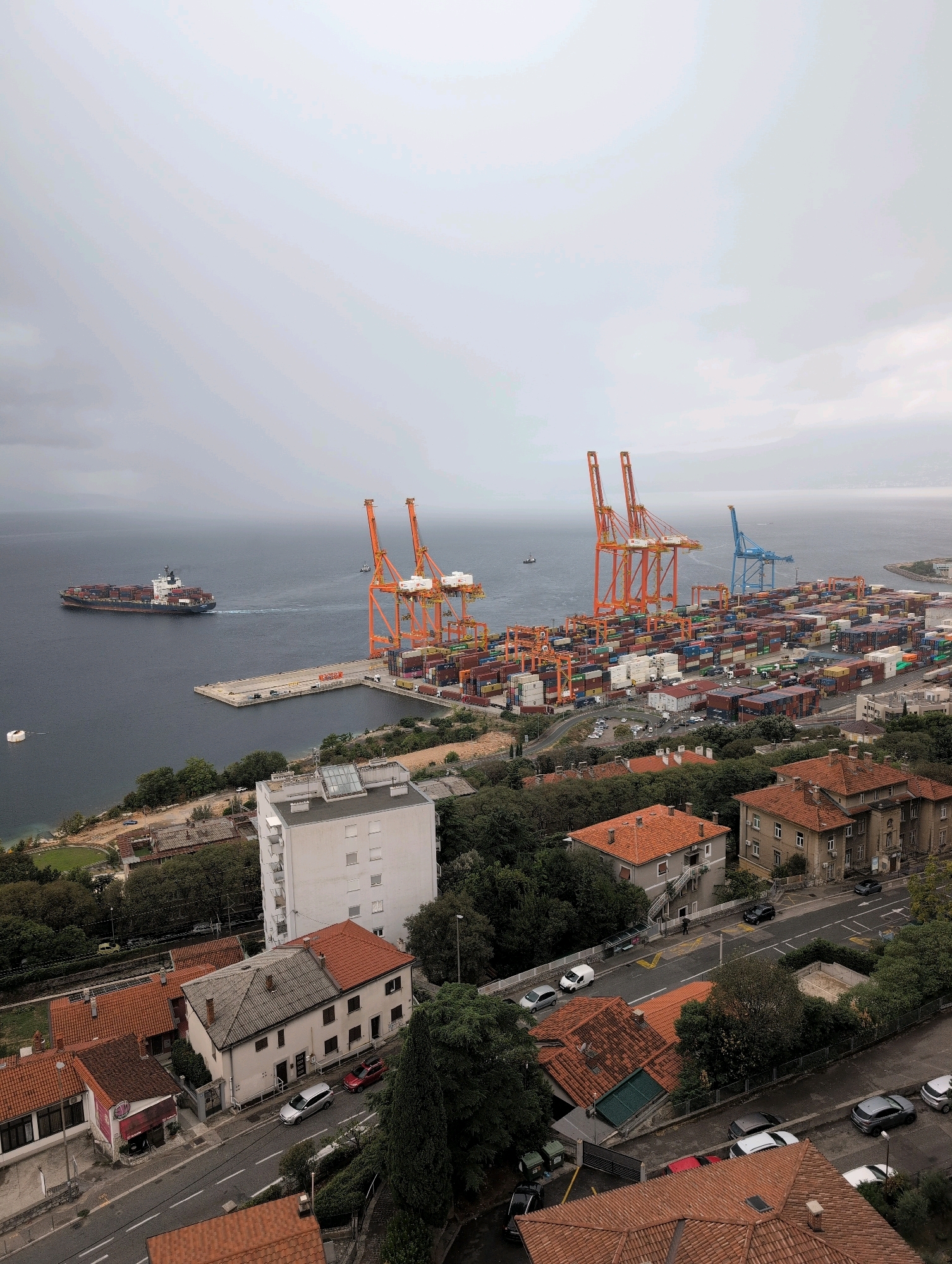An aerial, high-angle, full shot captures a port scene in Rijeka, Croatia. The sky is overcast, with a predominantly gray hue. In the foreground, residential buildings with red-tiled roofs are visible. The middle ground features more buildings and a winding road with parked cars. The background is dominated by a large container port with numerous colorful shipping containers stacked high and several large cranes, two of which are bright orange and one blue. A large cargo ship is sailing on the dark blue water to the left of the port.