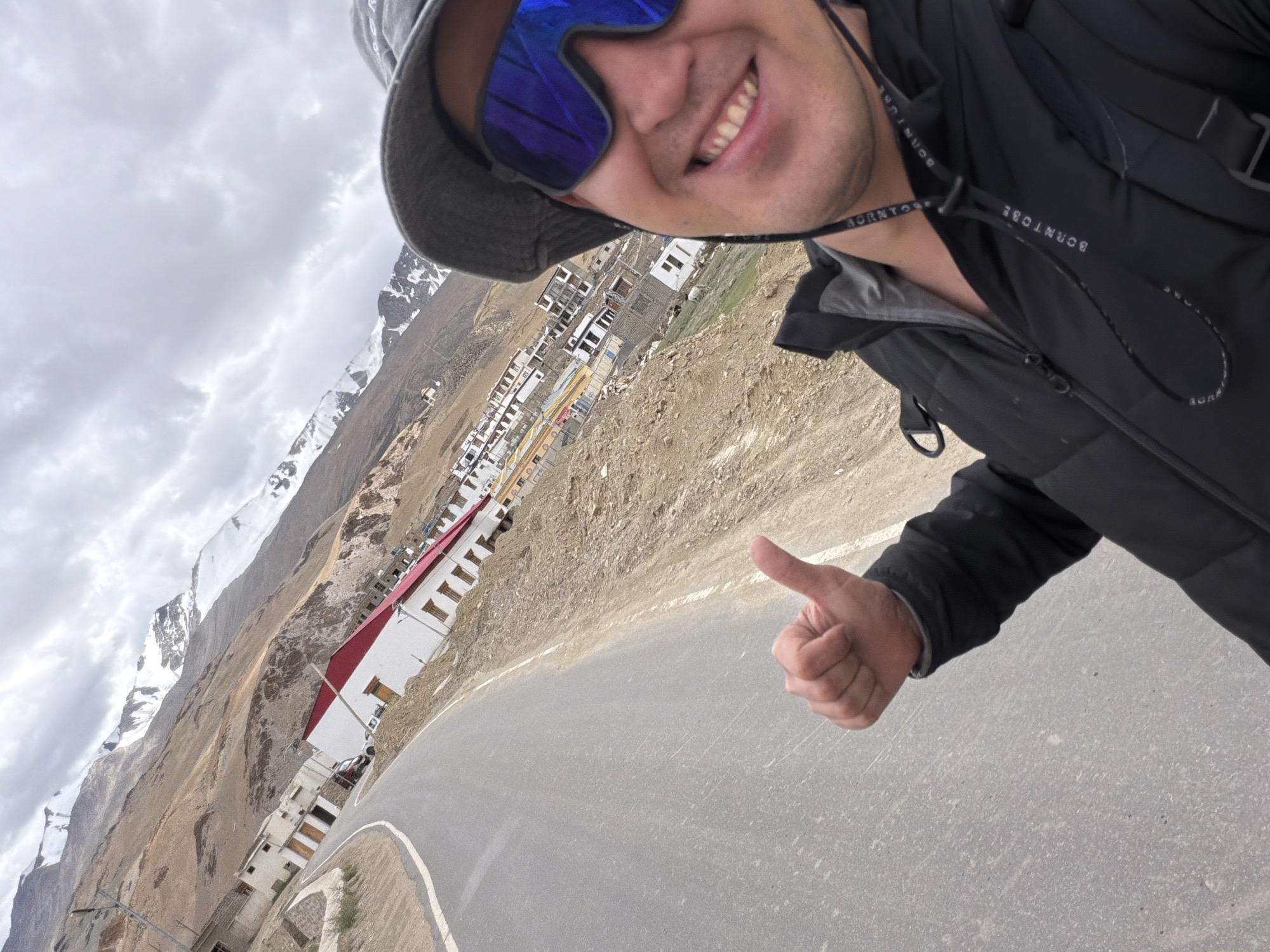 A first-person perspective selfie shows a man with a dark bucket hat and reflective blue goggles smiling widely. He is wearing a black puffer jacket with a gray inner layer and a black lanyard with white text that reads "BORNTODBE." His right hand is raised, giving a thumbs-up gesture. The man is positioned in the foreground against a backdrop of a steep, mountainous landscape. A paved road curves through the scene, leading towards a village of white buildings with red roofs. The mountains in the distance are snow-capped under a cloudy sky. The village is identified as being in Karzok, India. The environment is dry and arid, with rocky terrain and scattered vegetation.