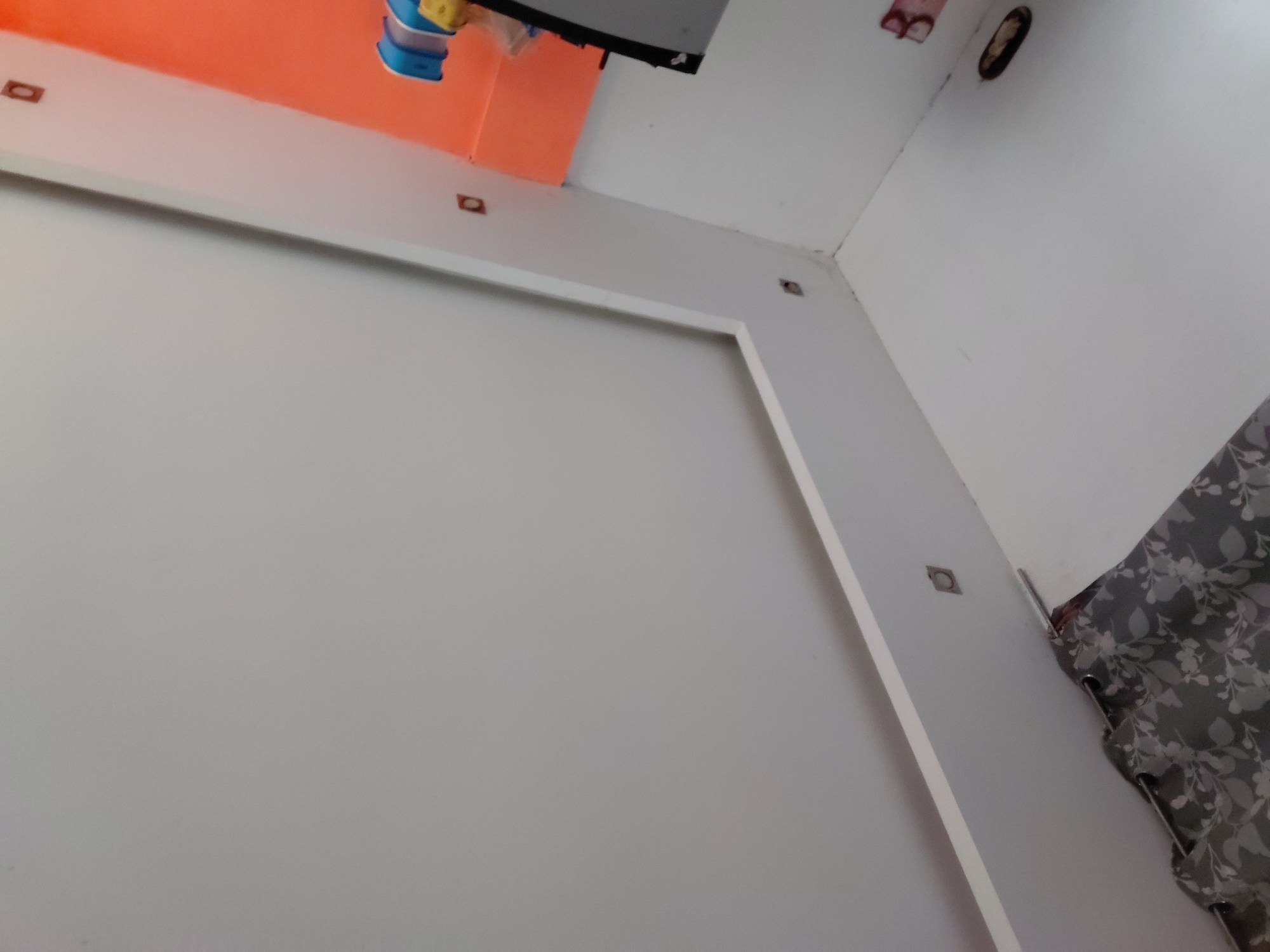 This upward-angled image captures an interior room in Santa Ana, Venezuela, showcasing white walls and ceiling with decorative elements. A large white rectangular molding outlines a section of the main wall, with small, square, dark insets visible along its length, possibly indicating recessed lighting or decorative accents. Similar square insets are also seen on the adjacent white wall.
Towards the upper left, an orange-painted wall or architectural feature contrasts with the white. Above this orange section, a black, flat object, potentially a screen or monitor, is partially visible, along with some blue and yellow objects that appear to be household items or containers. The white ceiling features a dark, oval-shaped opening or fixture. To the right, a grey curtain with a faint, lighter floral or leaf pattern is partially visible, drawn across what might be a window. No people are present, and no specific activity is depicted, providing a static view of the room's decor and fixtures.