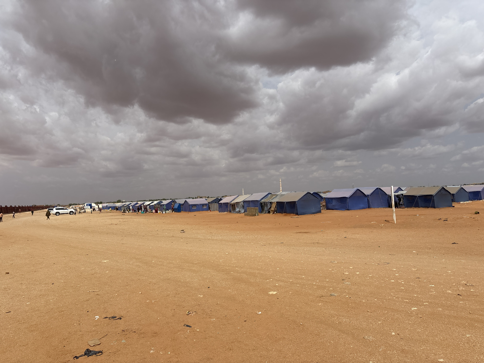 This image depicts a refugee camp in El-Obeid, Sudan, under a cloudy sky. The foreground is a vast expanse of dry, sandy ground littered with small debris. In the midground, a long row of blue tents, likely temporary shelters, stretches across the frame. Some tents are open, revealing their interiors or the people inside, while others are closed. Several people are visible walking around the camp, some interacting with each other or engaged in activities. A white SUV is parked near the tents, and other vehicles are also present in the distance. In the background, more tents and structures can be seen, suggesting a large settlement. The overall environment is arid and appears to be a functional, albeit basic, living space for the displaced population.