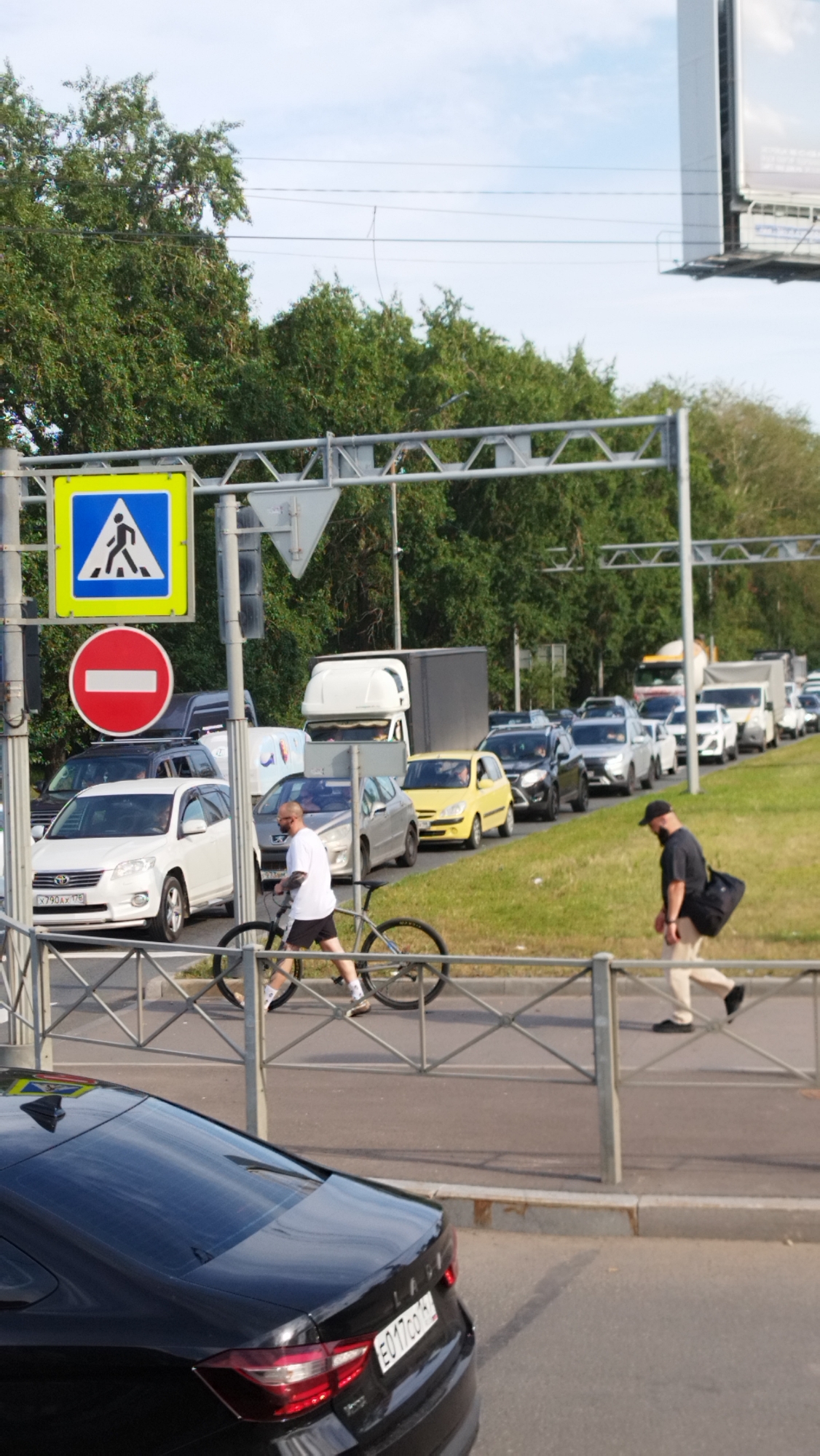 This image captures a busy street scene in Saint Petersburg, Russia, likely during the daytime with clear skies and sunshine. The primary focus is a traffic jam on a multi-lane road, with numerous cars and trucks of various colors and models forming long queues. The scene is set against a backdrop of lush green trees and a partly cloudy sky.
In the foreground, a pedestrian crossing sign is visible, indicating a designated area for people to cross the road. Nearby, a red circular sign with a horizontal white bar, signifying "no entry" or a prohibition, is also present.
Two men are walking near the pedestrian crossing. One man, wearing a white t-shirt and black shorts, is pushing a bicycle. He appears to be casually walking, possibly navigating the traffic or heading towards his destination. The other man, dressed in a dark t-shirt and light-colored pants, carries a black bag over his shoulder and walks with a focused stride.
In the lower part of the image, the rear of a black Lada car is visible, its license plate reading "E017co 61". The car is parked or moving slowly within the traffic flow.
The overall impression is one of urban congestion, common in large cities, with everyday activities like cycling and walking taking place alongside the stalled traffic. The presence of specific road signs and the car's license plate offer clear clues to the location.