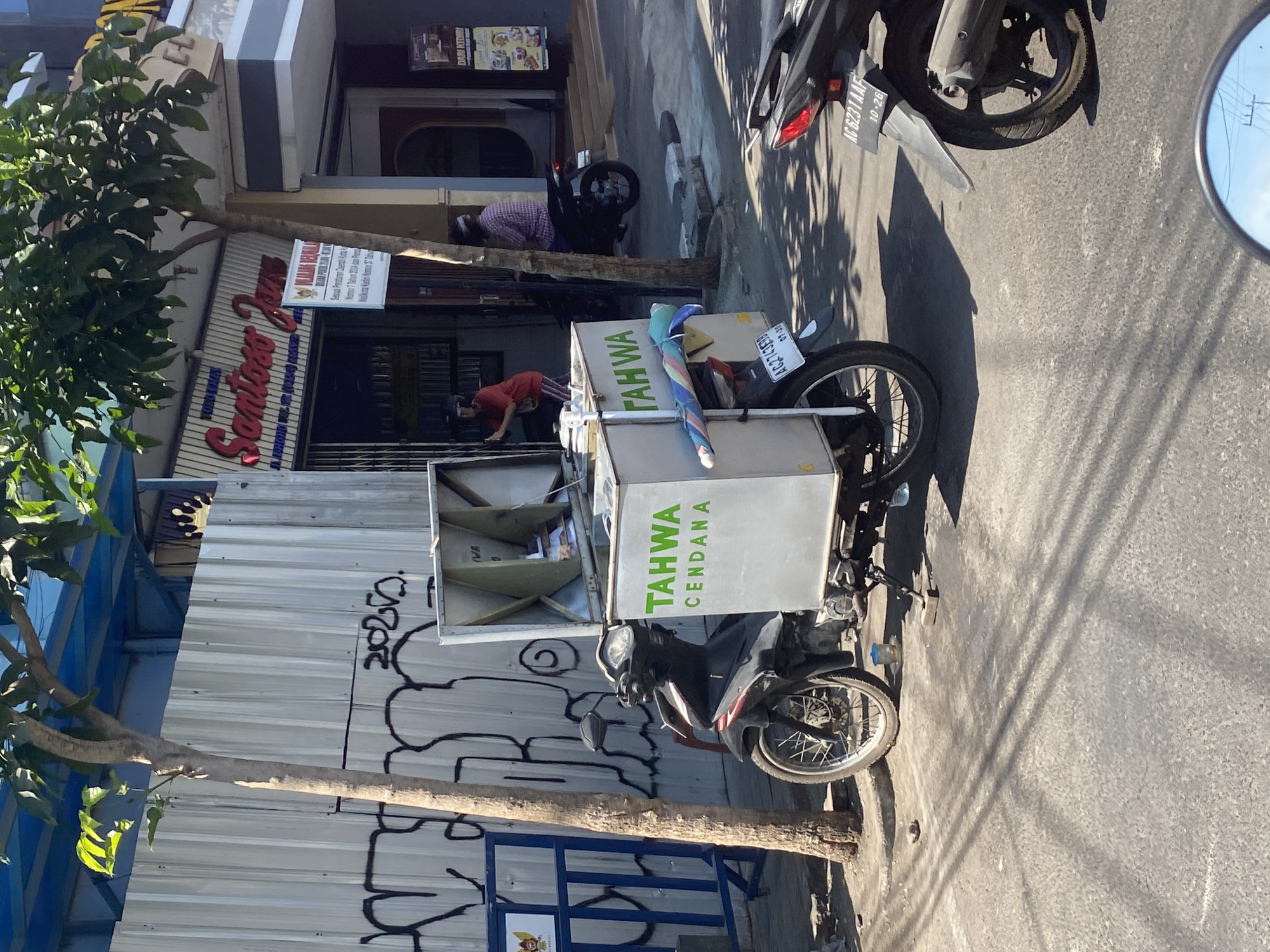 This image, taken in Kediri City, Indonesia, depicts a street scene with a focus on a motorcycle equipped with a large cargo box. The motorcycle, a dark color with red accents, is parked on the asphalt road, facing towards the right of the frame. The cargo box is white with green lettering that reads "TAHWA CENDANA". A person in a red shirt and striped pants is visible near the open cargo box, appearing to be interacting with its contents.
Behind the motorcycle is a corrugated metal wall covered in black graffiti. To the right of the wall is a storefront with a blue awning and a sign that reads "Santoso Jaya". In front of the storefront, a person in a plaid shirt is seated on another dark motorcycle, facing away from the camera. A second motorcycle, partially visible at the bottom right of the frame, also has a license plate. The environment is sunny with strong shadows cast by trees and objects. The street appears to be a commercial area with buildings and sidewalks.