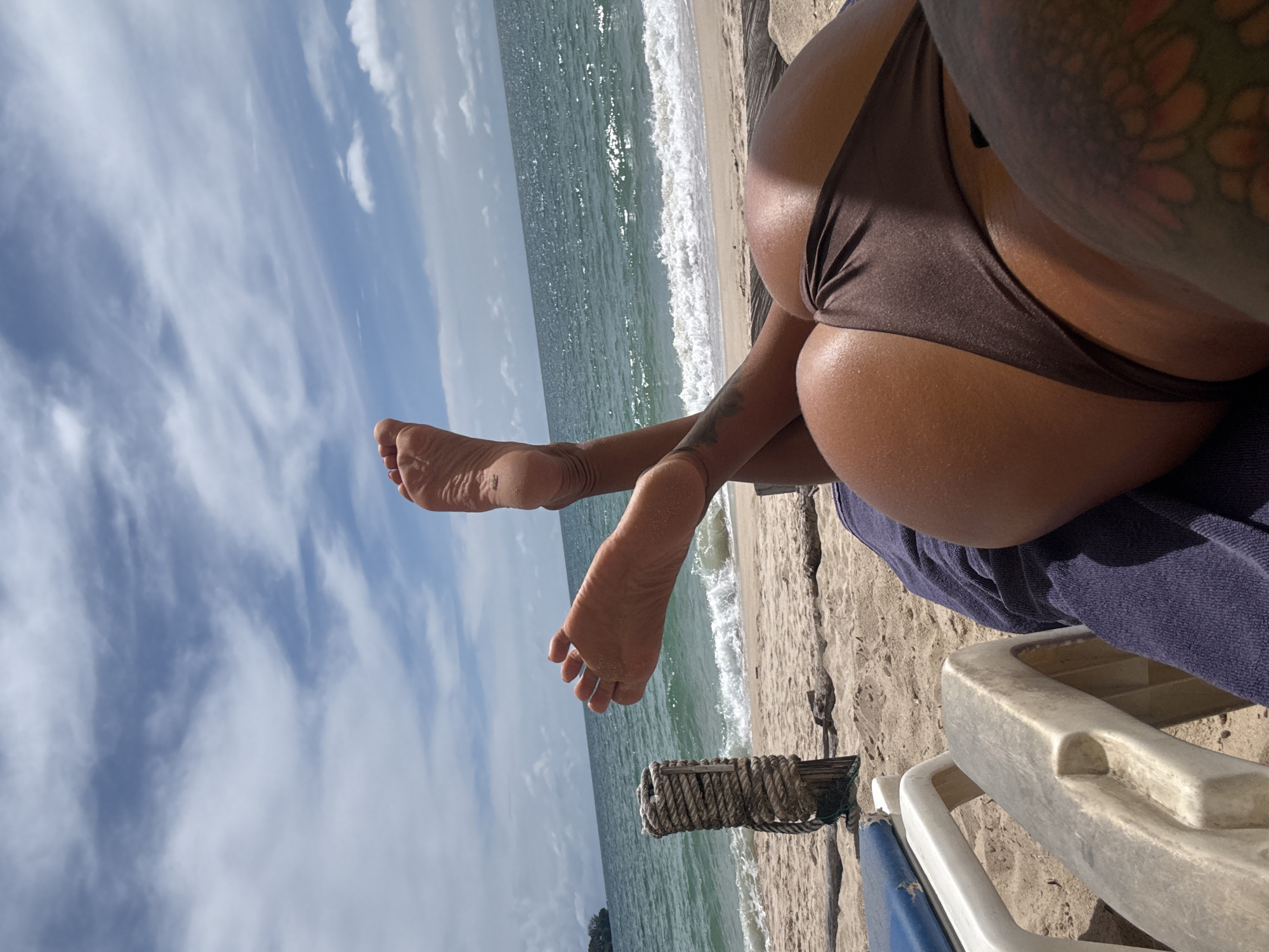 A person lies on a beach chair on the sand, facing away from the camera, with their legs extended towards the ocean. The person is wearing a brown bikini bottom. Their feet are raised and visible, with toes spread. The ocean and a cloudy sky are in the background. A wooden post with rope is to the left of the person.