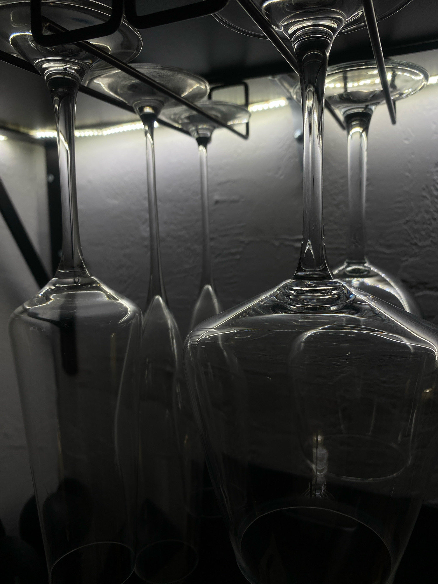 This image is a close-up, low-angle shot of several empty wine glasses hanging upside down from a metal rack. The glasses are made of clear glass and have long stems. The rack appears to be mounted on a wall, as a textured surface is visible in the background. A strip of LED lights runs along the underside of the rack, casting a bright glow upwards and creating reflections on the glasses. The lighting is dramatic, with strong contrasts between light and shadow, highlighting the curved surfaces of the glass. There are no people or animals visible in the image. The scene is indoors, likely in a kitchen or dining area, possibly in a home or establishment in Zarechny, Russia, given the prompt's context, although the visual elements do not explicitly confirm this location.