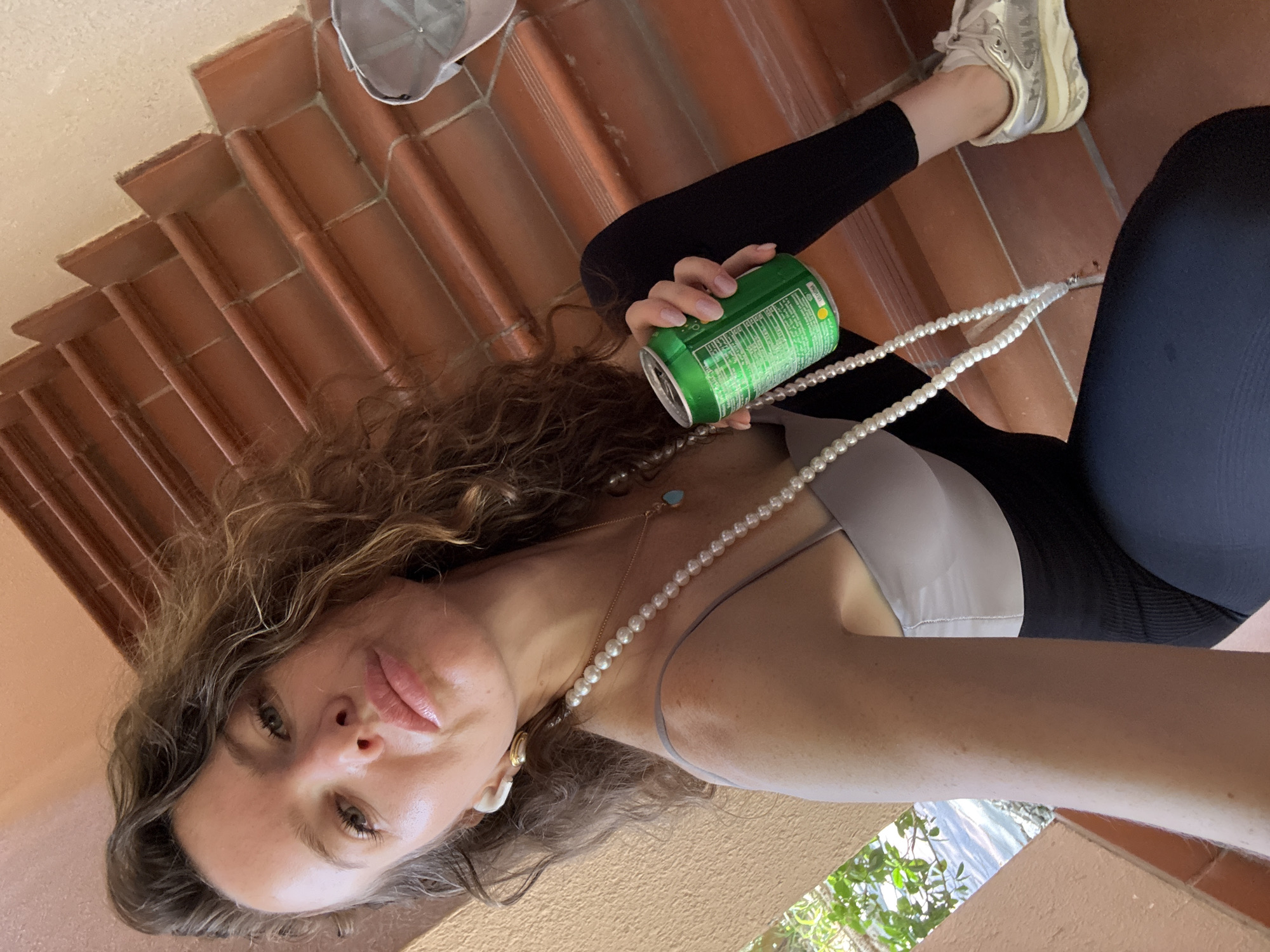 The image shows a woman posing for a selfie on a terracotta-tiled staircase, likely outdoors given the visible foliage and water in the background. The photo is taken from a low angle, looking upwards at the woman.
**Subjects:** The main subject is a woman with long, wavy brown hair. She is wearing a light-colored (possibly white or light grey) sleeveless top, black leggings, and light-colored sneakers with a thick sole. She has light-colored pearl earrings, a long pearl necklace, and a delicate gold chain necklace with a small turquoise pendant. She is holding a green can with a white label in her right hand.
**Scene and Setting:** The woman is seated on a terracotta-tiled staircase. To her right, there's a glimpse of green foliage and a body of water, suggesting a coastal or waterfront location. The tiles appear to be in good condition, and a light-colored cap is visible resting on one of the steps behind her.
**Activity:** The woman appears to be taking a selfie, holding the camera or phone at arm's length. Her gaze is directed towards the camera, and her lips are slightly parted. She is holding a beverage can.
**Notable Details:**
* **Time of Day/Weather:** The lighting suggests daytime, possibly bright sunshine, though no harsh shadows are visible.
* **Location Cues:** The terracotta tiles and the glimpse of water and foliage hint at a warm, possibly Mediterranean or tropical environment. Given the provided context of Dubai, United Arab Emirates, this would align with a sunny, outdoor setting.
* **Emotions:** The woman's expression is neutral to slightly contemplative, with a subtle pout.
* **Visible Text:** The green can she is holding has white text on its label, but it is not clear enough to be read.
The image captures a casual moment, possibly during a break from an activity, with the woman enjoying a drink and taking a picture.