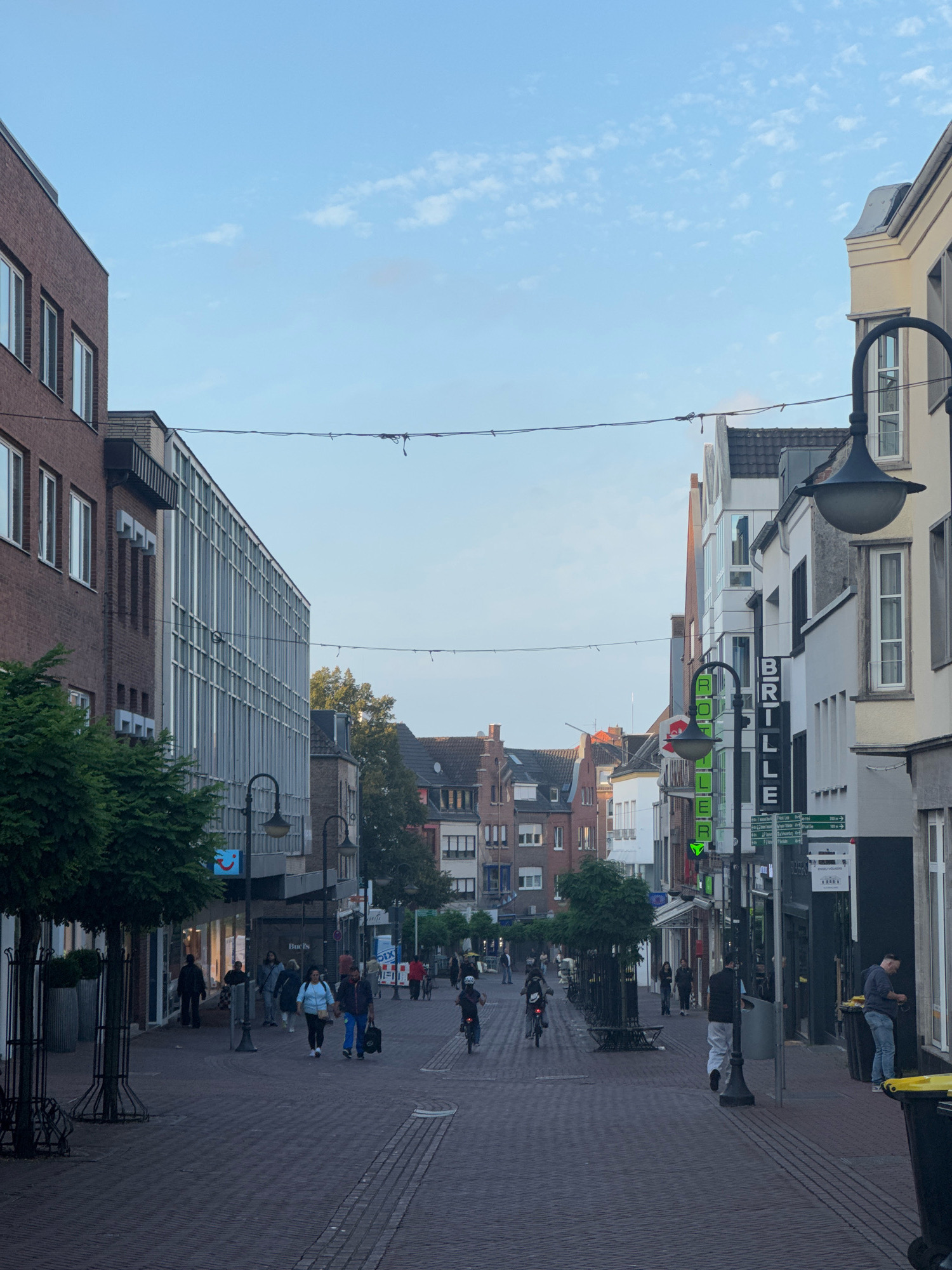 The image depicts a street scene in Cleves, Germany, characterized by a pedestrian walkway paved with red brick. The walkway is lined with buildings on both sides, featuring a mix of architectural styles. On the left, a modern building with a glass facade stands next to a brick building. On the right, a series of multi-story buildings display various shopfronts and architectural elements, including a sign that reads "BRILLE".
Several individuals are present in the scene, engaged in various activities. Some are walking along the street, while others are seen riding bicycles. The people appear to be a mix of ages and genders, dressed in casual attire. Objects such as streetlights, trees in planters, trash receptacles, and shop signage are visible throughout the environment. The sky above is blue with scattered white clouds, indicating daytime. The overall atmosphere suggests a typical urban street environment during daylight hours.