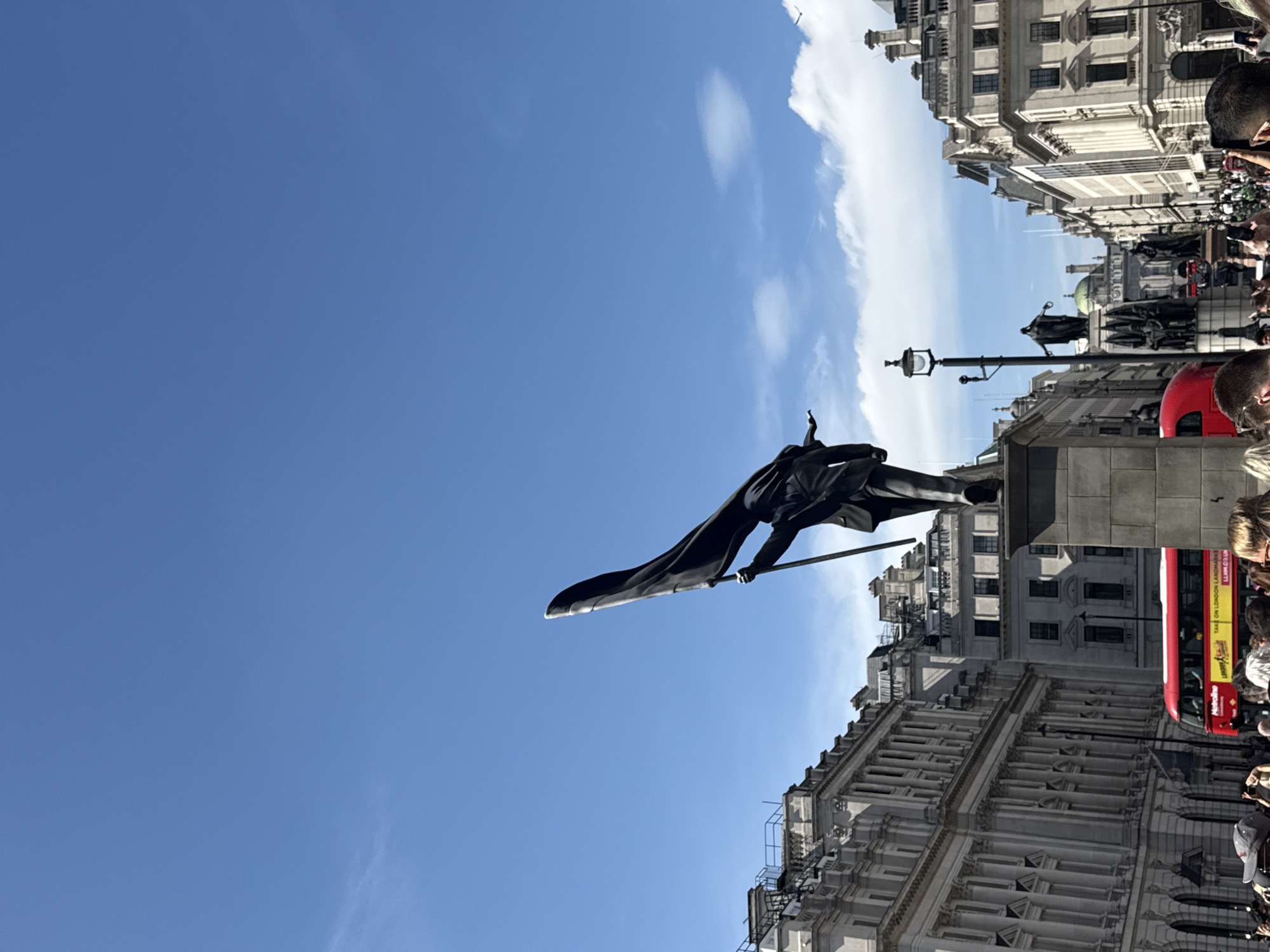 The image shows a view of Trafalgar Square in London, United Kingdom. The statue of King Charles I is prominently featured in the foreground. Buildings with classical architectural details line the square. A double-decker red bus and crowds of people are visible in the background. The sky is blue with some clouds.