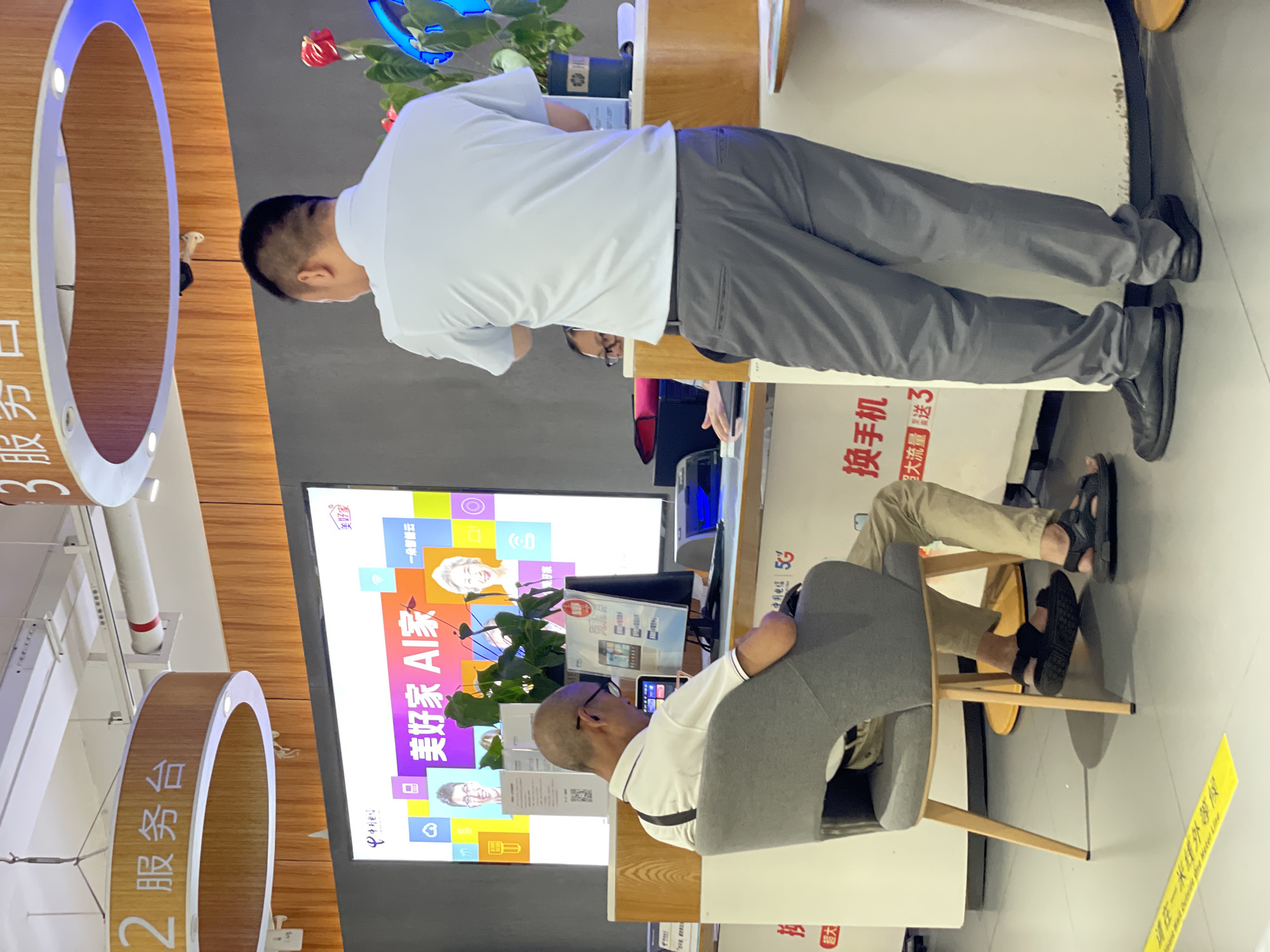 The image depicts an indoor scene, likely a service counter in a telecommunications or technology store, possibly in Nan'an District, China, given the presence of Chinese text. In the foreground, a man is seated on a grey chair, facing away from the camera and looking at a computer monitor. He is wearing a white polo shirt, beige pants, and black sandals. To his right, another man stands facing the counter, wearing a light blue short-sleeved shirt and grey pants. He appears to be interacting with someone behind the counter, though only a part of their face is visible. Behind the seated man, a large screen displays graphics and text, including what appears to be "美好家 AI家" which translates to "Beautiful Home AI Home." Various promotional posters are visible on the counter and walls, with Chinese characters indicating services like "换手机" (phone exchange) and "5G". The environment appears to be well-lit with fluorescent ceiling lights and decorative circular wooden light fixtures overhead labeled with numbers. A potted plant with red flowers is visible on the counter. The floor is tiled and a yellow line with Chinese text is marked on it.