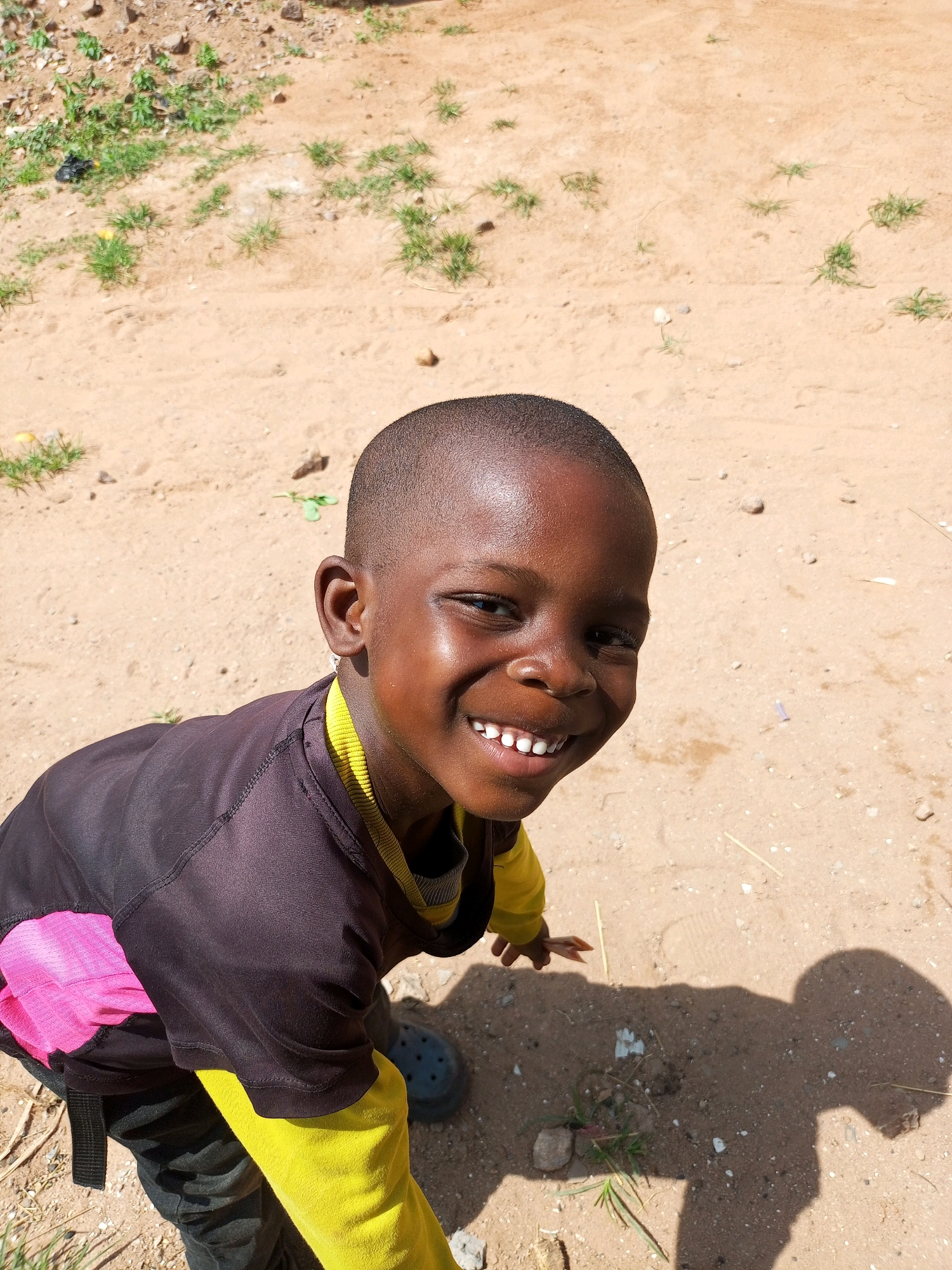 A young Black boy, with a close-cropped haircut and dark skin, smiles broadly at the camera. His teeth are white and perfectly aligned. He wears a black long-sleeved shirt over a bright yellow long-sleeved undershirt. A bright pink pouch is attached to his clothing on the left side. He is wearing dark pants and dark blue Croc-style shoes. The boy is outdoors in what appears to be a dusty, sandy area with patches of dry grass and scattered rocks. The ground is a light tan color. The boy's shadow is cast on the ground to his right. The image was taken from a slightly elevated perspective, looking down at the boy. The overall impression is one of joyful innocence in a simple, outdoor setting. The location is Tempure, Nigeria.