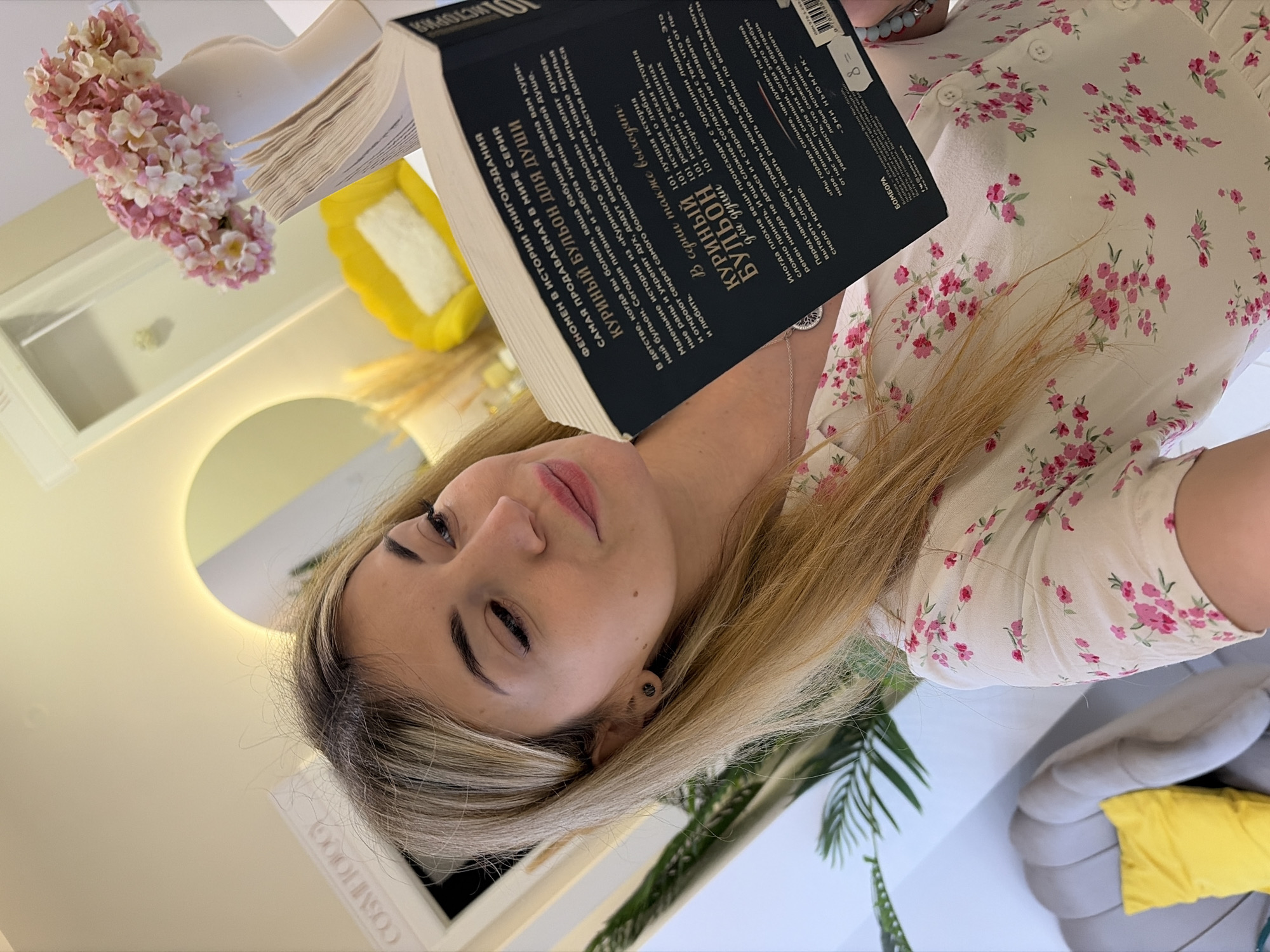 A young woman is holding a book titled "Chicken Soup for the Soul." The book is in Russian, and the text on the cover suggests it is a collection of stories for the soul, promoting sharing and love. She has fair skin, blonde hair with darker roots, and is wearing a light-colored floral-patterned shirt. She has a stud earring in her left ear.
The environment includes a white surface, possibly a table or counter, with pink and white flowers in a vase to the left. There is a yellow pillow and a gray sofa cushion in the background. A circular mirror is reflecting the surroundings, and a plant is visible in the lower right corner.
The setting appears to be indoors, possibly a cafe or a home interior. There is no indication of the city of Budva, Montenegro.