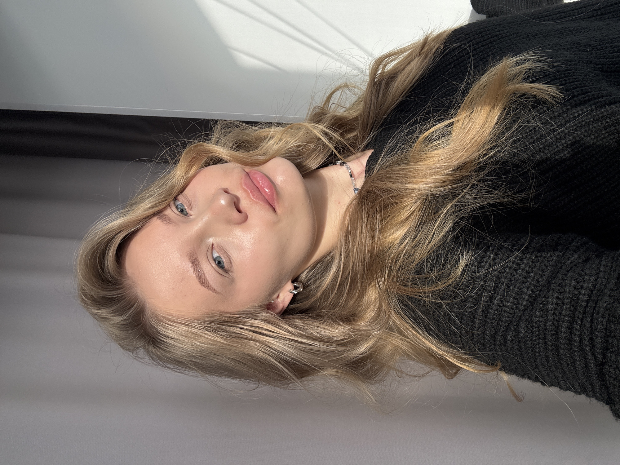This is a close-up portrait of a young woman, photographed in Chaikovsky, Russia. The image is oriented sideways, showing her head and upper torso from a slightly low angle, as if she is lying down and looking up. She has long, wavy blonde hair that cascades around her shoulders and across her black knitted sweater. Her light blue eyes are open, looking towards the viewer, and her lips are slightly parted, adorned with a subtle pink lip gloss. Her eyebrows are well-defined. She wears a delicate silver-toned bracelet on her wrist with a small, possibly blue or clear, charm, and a silver ear cuff or earring is visible on her left ear. The background features plain light-colored surfaces, with strong, distinct shadows suggesting natural daylight streaming in through a window, possibly filtered by blinds or slats, indicating a sunny day. The lighting is bright, highlighting her features and the texture of her hair. The overall impression is calm and serene, capturing a posed yet natural moment. No text is visible in the image.