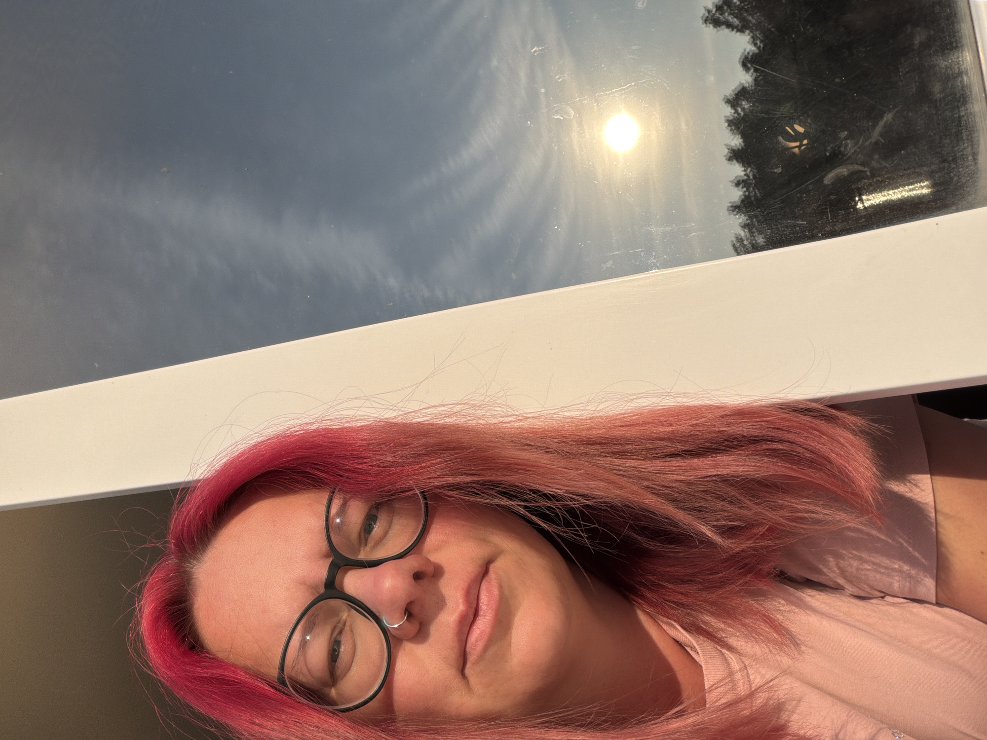 A selfie taken by a woman with bright pink hair and glasses shows her face and upper torso. She is wearing a pink t-shirt. She is looking directly at the camera with a neutral expression. A white vertical beam is in the foreground, slightly to the right of the woman. To the right of the beam, a window shows a bright sun low in a blue sky with some wispy clouds. The reflection in the window shows a dark silhouette of trees. This image was taken outdoors in Chaikovsky, Russia, during daylight hours.