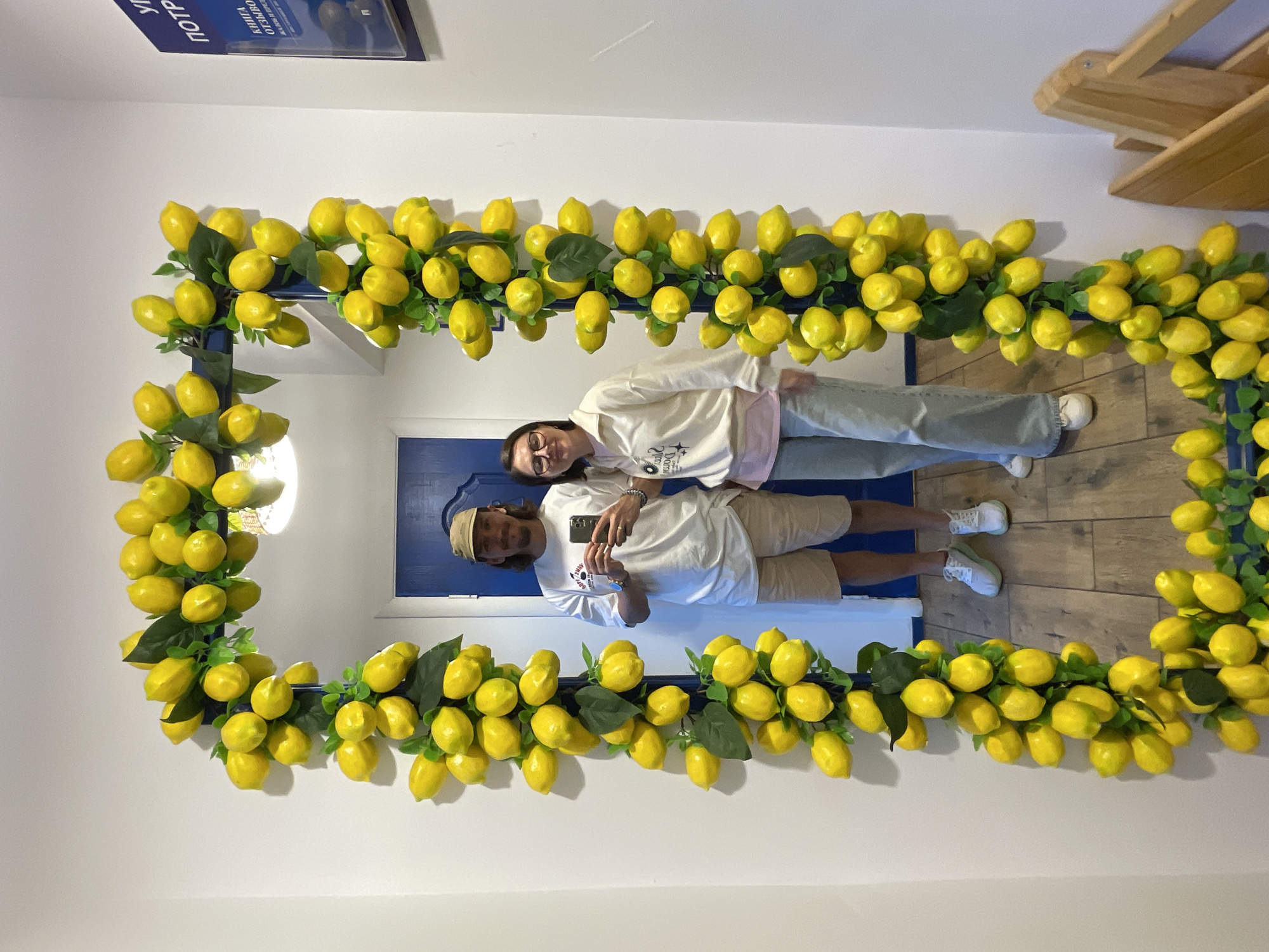 The image shows a couple taking a selfie in front of a lemon-themed mirror. The mirror frame is heavily decorated with artificial lemons and green leaves. The couple is standing close together, holding a smartphone to capture the photo. The man is wearing a light-colored t-shirt, khaki shorts, and a hat, while the woman is wearing a white sweatshirt and light-colored jeans. The reflection shows a blue door behind them and a wooden floor. The location appears to be an interior space, possibly a shop or a decorated room. There is no immediate evidence to suggest the image was taken in Izhevsk, Russia.