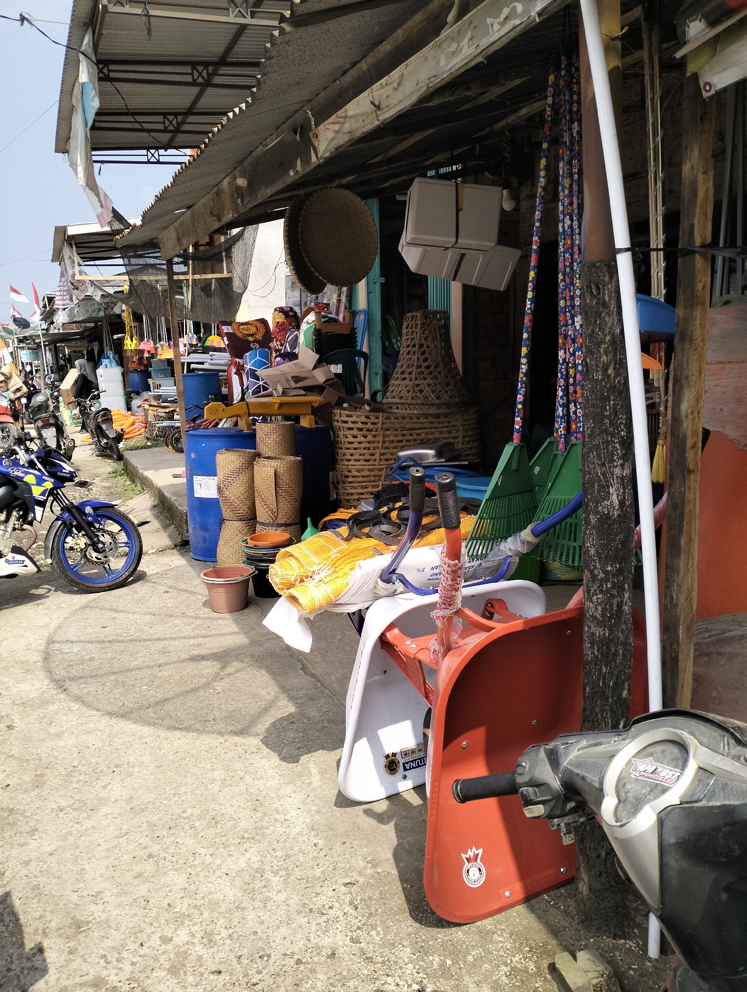The image depicts a street market scene in Serdang Jaya, Indonesia. The foreground features a portion of a black and silver motorcycle, its handlebars and instrument panel visible. To the left and center, various items are displayed on the concrete ground and on a raised curb. These include stacked woven baskets, blue plastic barrels, and what appear to be rolled-up mats or fabric in yellow and white. Behind these, a red plastic chair with a white seat is prominent. Several green leaf rakes with patterned handles are leaning against a wooden post. In the background, stalls with corrugated metal roofs are visible, displaying an assortment of goods such as brooms, bags, and other household items. A few motorcycles are parked along the curb. The overall impression is one of a bustling, open-air market with goods laid out for sale under a covered walkway.