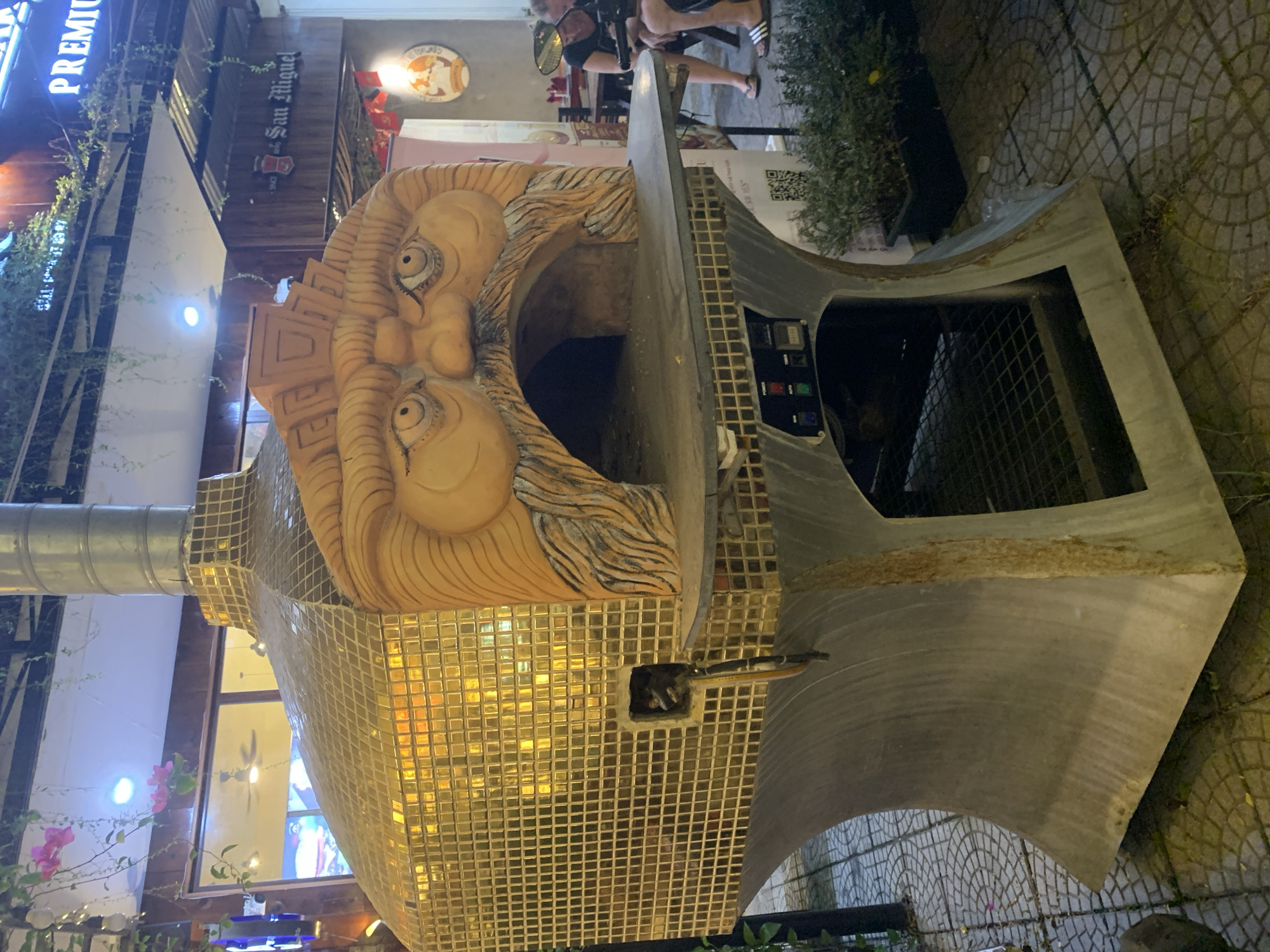 The image shows a unique, gold-tiled pizza oven with a large, carved face design above the opening. The oven is situated on a tiled outdoor patio, likely in Đà Nẵng, Vietnam, given the context of similar establishments. The oven has a metallic chimney and a grey concrete base with visible controls and a shelving unit. In the background, there are people seated at tables, suggesting a restaurant or dining area. A sign for "San Miguel" is visible, along with some greenery and string lights overhead. The overall scene depicts an outdoor dining or food preparation area with a distinctive, artistic pizza oven as the focal point.