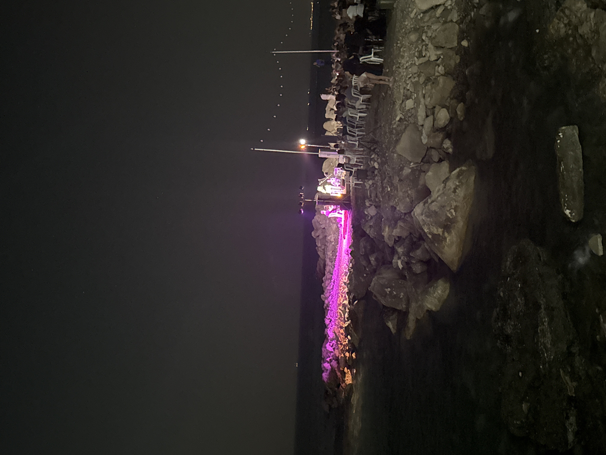 An outdoor concert is taking place at night on a rocky shore. A stage is set up with purple and pink lighting, and a band is performing. An audience of people is seated in chairs facing the stage. The concert is held in Agios Tychona Community, Cyprus, along the Mediterranean Sea. The sky is dark, and the water is calm. The scene suggests a festive and lively atmosphere.