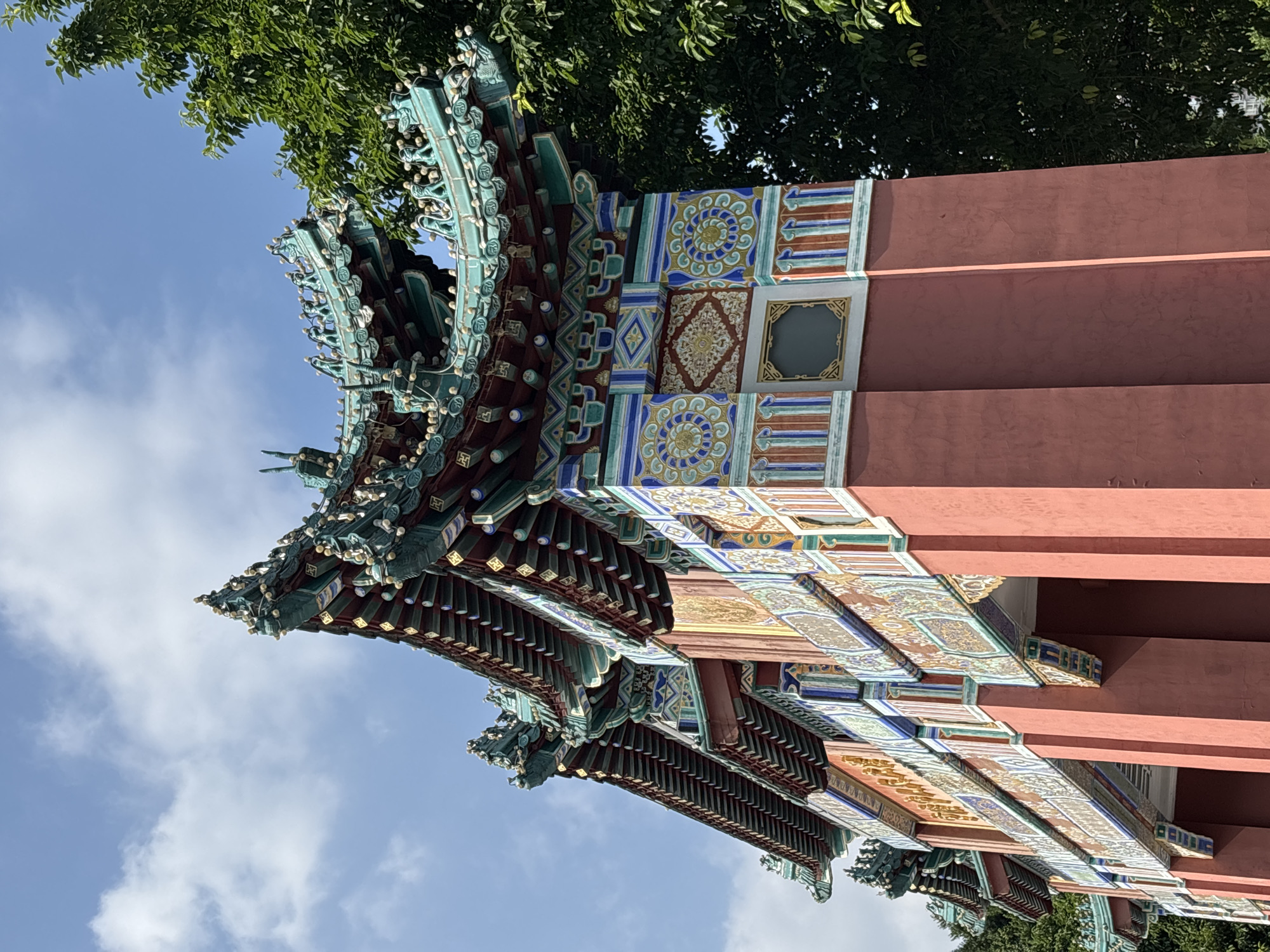 A low-angle outdoor photograph captures the ornate roof and upper facade of a traditional Chinese archway in Yuzhong District, China. The archway features a multi-tiered, curved roof adorned with intricate green ceramic tiles and decorative figures, possibly mythical creatures, along its edges. The supporting pillars and upper walls are painted a vibrant coral red and decorated with colorful, geometric patterns in blue, yellow, and white. These patterns include circular motifs and intricate scrollwork. A large, lush green tree is visible to the right of the archway, partially obscuring the sky. The sky is a clear blue with scattered white clouds. The overall scene conveys a sense of grandeur and architectural splendor.