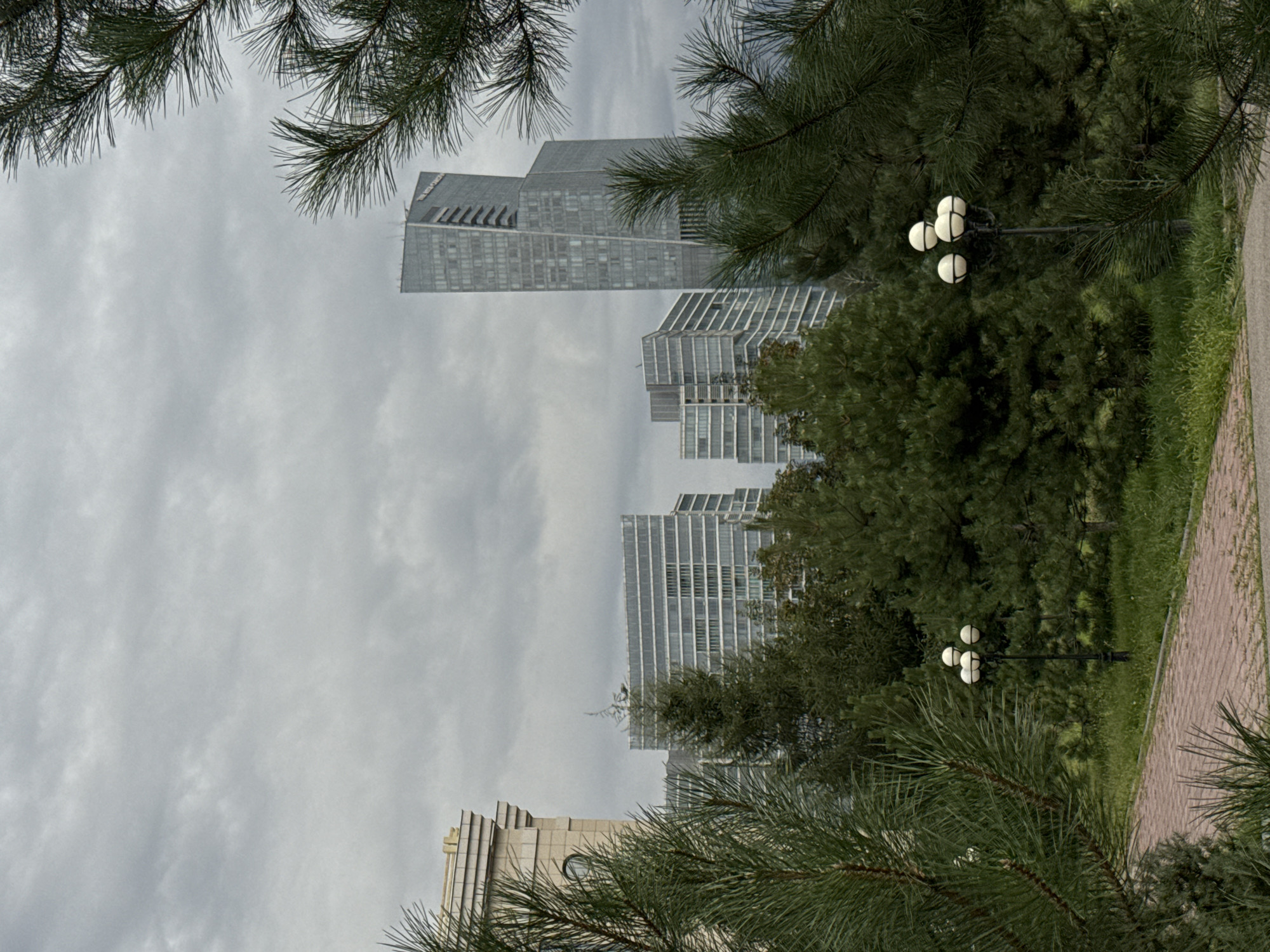 A medium shot captures a grey, overcast day in Almaty, Kazakhstan, featuring a blend of urban architecture and lush greenery. In the background, a modern, sleek skyscraper with a unique angled design dominates the skyline, flanked by other contemporary buildings constructed with glass and metal. In the foreground, dense coniferous trees form a natural barrier, partially obscuring the buildings. A paved pathway, constructed of red bricks, winds through the park-like setting, with green grass and bushes dotting the landscape. A black lamppost with four spherical lights stands near the pathway, adding a touch of artificial illumination to the scene. The overall atmosphere is serene and understated, with the muted colors and diffused light contributing to a sense of calm.