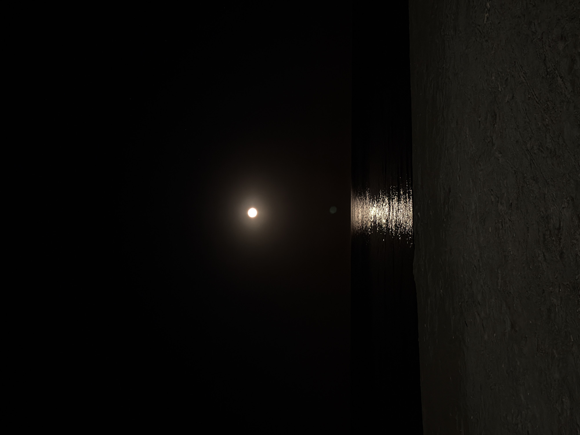 The image depicts a nighttime scene, likely in Larnaca, Cyprus, given the location context. The primary subject is the moon, appearing as a bright, luminous orb in a dark sky. Its reflection shimmers on the surface of dark water, creating a trail of light that stretches towards the right side of the frame. The water is calm, with only the reflection indicating its presence.
To the right of the water, a dark, textured surface occupies the remainder of the image. This surface appears to be a rough, possibly stone or concrete wall, with visible irregularities and shadows that emphasize its texture. The contrast between the smooth, reflective water and the rough, dark wall is stark.
The scene evokes a sense of tranquility and mystery, characteristic of a moonlit night. The absence of people or man-made structures, apart from the implied presence of the wall, contributes to the serene and isolated atmosphere. The time of day is clearly night, indicated by the darkness and the presence of the moon. No specific weather conditions are apparent, other than the calm water. There is no visible text in the image.