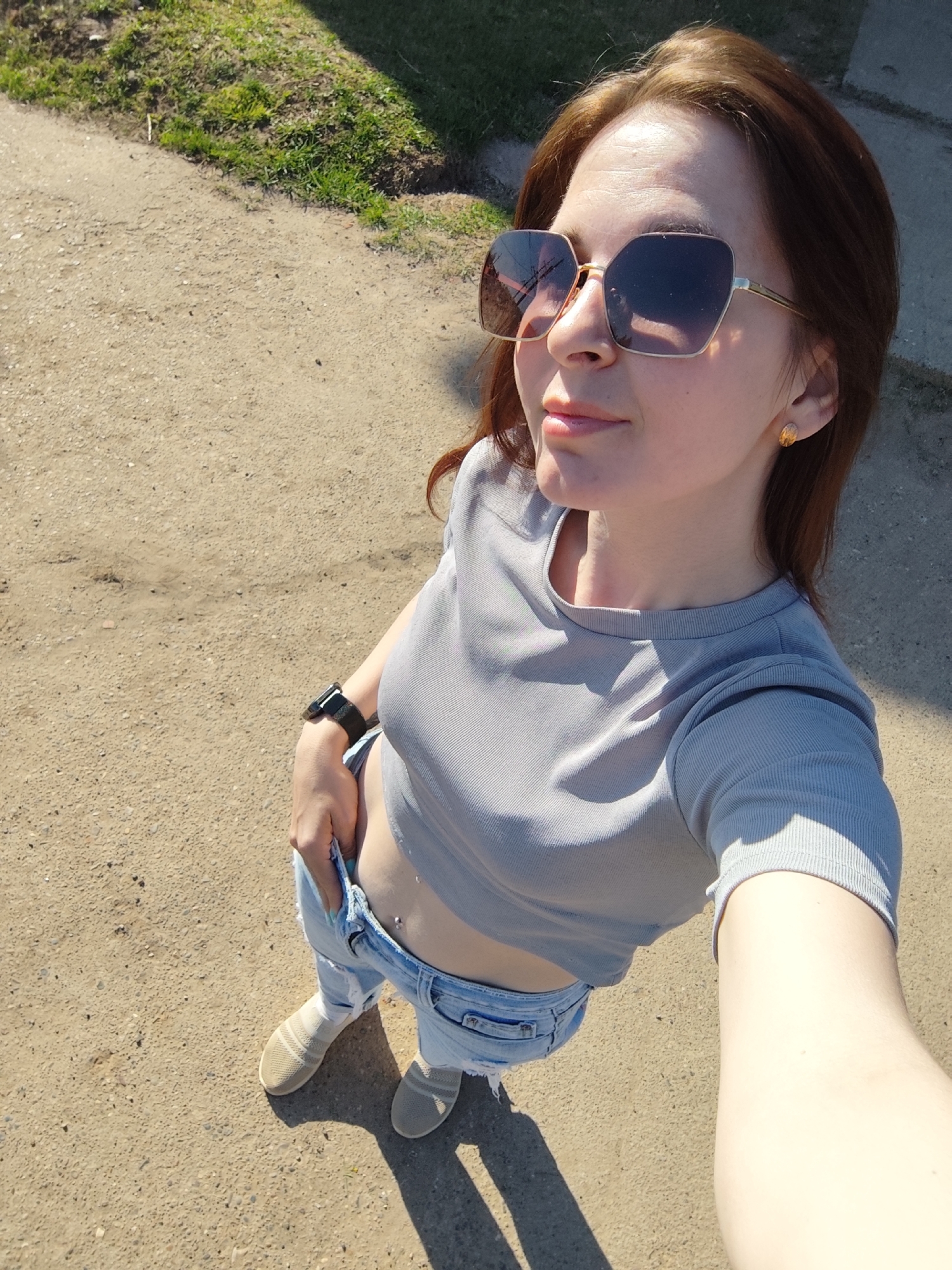 A woman is standing outdoors on a paved or dirt path with sparse grass visible near the edge. She is wearing sunglasses, a grey short-sleeved crop top, light blue jeans, and beige slip-on shoes. She has a watch on her left wrist, and her left hand is in the pocket of her jeans. The photo is taken from a low angle, looking up at the woman. The lighting suggests it is a sunny day. Based on location constraints, I could not pinpoint a specific street or landmark.