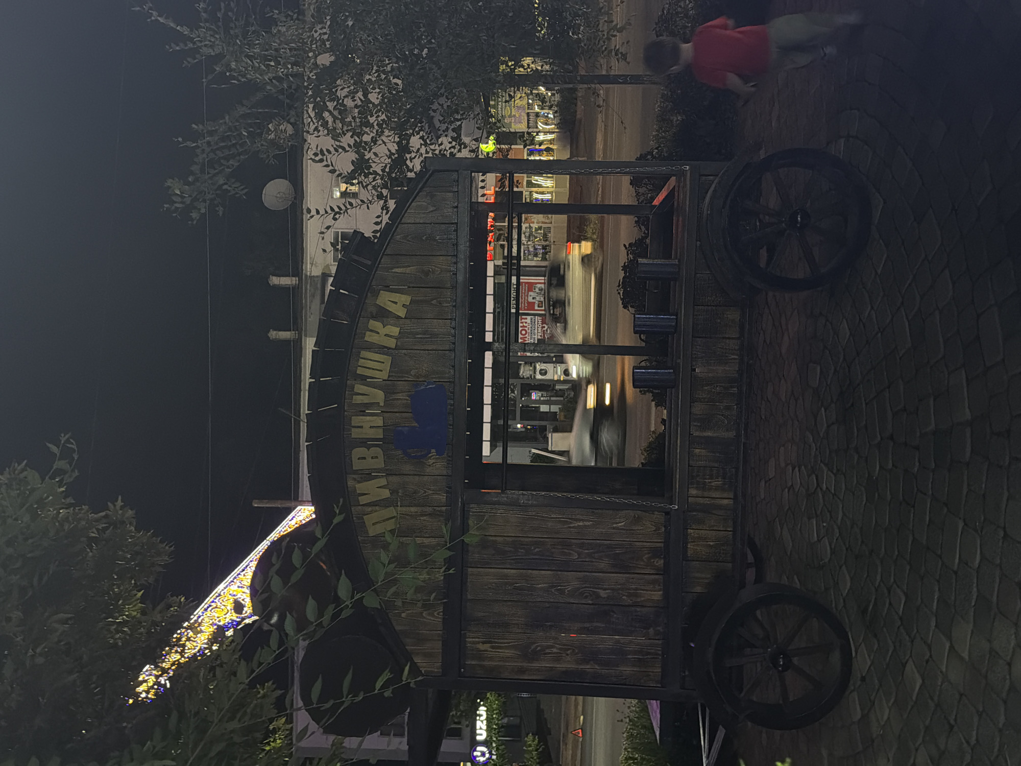 In the city of Chirchik, Uzbekistan, at night, a wooden food cart is parked on a cobblestone surface. The cart is branded with the Cyrillic word "ПИВНУШКА" (Pivnushka), meaning "beerhouse," and features a blue silhouette of a beer mug. In front of the cart, a young child wearing a red shirt and grey pants is running away from the camera. The background reveals a street with blurred car lights, storefronts with illuminated signs, and trees. A string of festive lights is visible in the upper left corner.