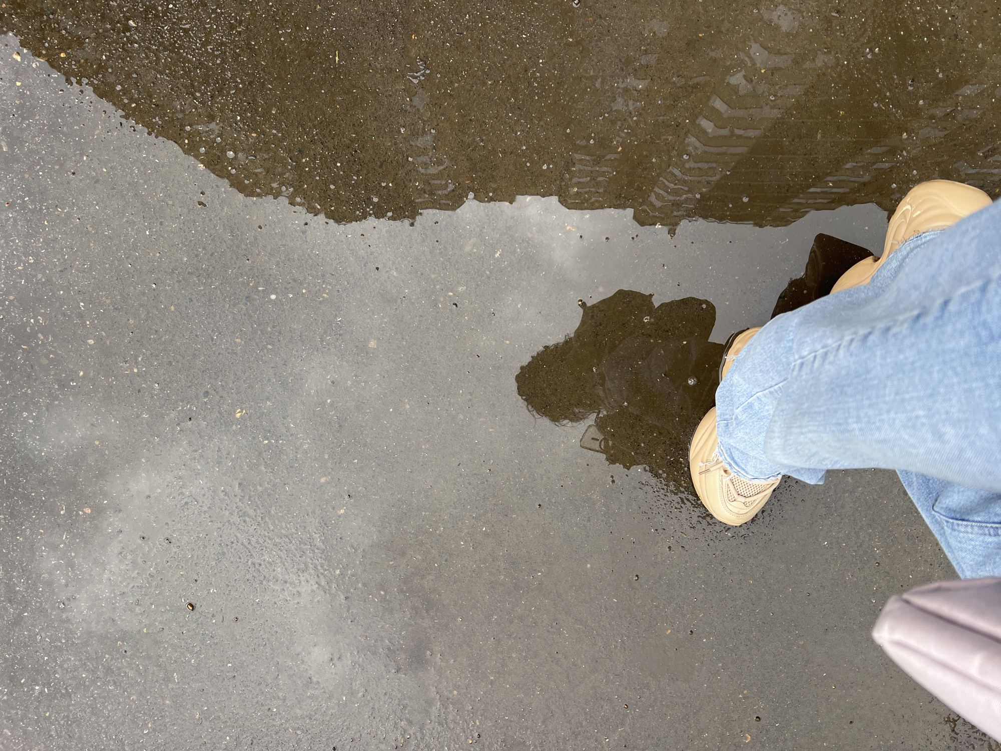 The photograph shows a puddle on a gray asphalt surface. A pair of light blue jeans and a beige sneaker are partially visible, suggesting a person is standing near the puddle. A faint shadow of a person and the reflection of a building with tire track marks are visible in the puddle. The location is suggested to be Krasnodar, Russia, although this is not directly verifiable from the image.