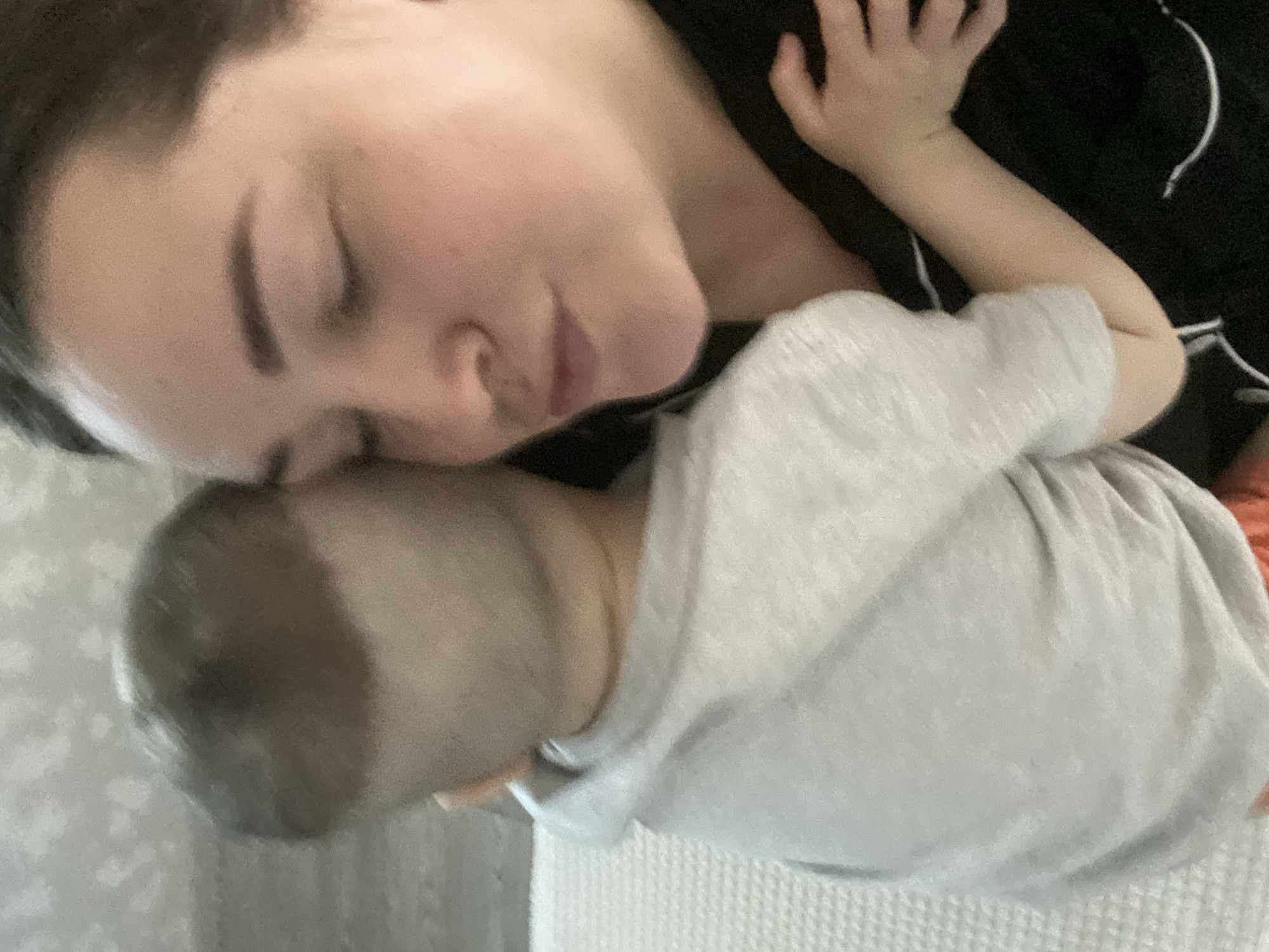 This close-up, slightly tilted photograph captures a tender and peaceful moment between an adult and a young child resting together indoors in Sochi, Russia. The adult, likely a woman, occupies the upper left portion of the frame, with her eyes closed and a serene expression on her face, suggesting she is sleeping or deeply relaxed. Her head is gently resting against the child, conveying a strong sense of intimacy and comfort. She has dark hair and is wearing a dark-colored top, possibly black, with a visible light-colored pattern or piping.
The young child is positioned below the adult, primarily showing the back of their head with light brown hair and their upper body. The child is wearing a light grey top. A small hand of the child is visible near the adult's face, adding to the affectionate nature of the interaction. The background is soft and indistinct, featuring light-colored surfaces that appear to be bedding or a blanket, with a subtle texture resembling a waffle weave visible towards the bottom right. The image evokes feelings of warmth, love, and tranquility. No specific time of day is discernible, and there is no visible text within the photograph.