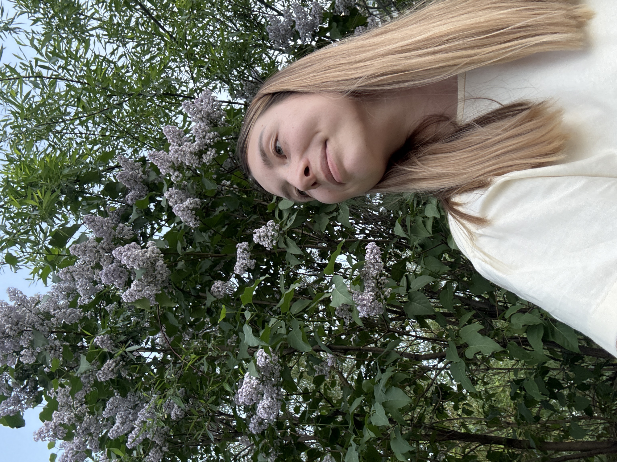 A woman with fair skin and blonde hair is in the foreground, angled towards the camera and slightly tilted to the side. She is wearing a light-colored shirt. Behind her is a blooming lilac bush with purple flowers and green leaves. The image appears to be taken outdoors during daylight. The location is possibly a park or garden in Moscow, Russia, inferred from the common presence of lilac bushes in urban green spaces there.