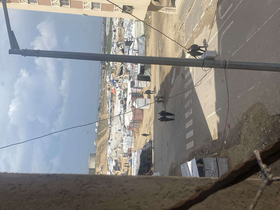 An elevated view captures a scene in Khan Yunis, Palestinian Territory, under a partly cloudy sky. In the immediate foreground, to the left, is the textured concrete edge of a building from which the image is taken. To the right, a dark grey utility pole with a streetlamp fixture and associated wiring stands vertically, obscuring a portion of a multi-story, beige-colored residential building with ornate brown window frames.
The midground features a paved asphalt road marked with a white pedestrian crosswalk. Six adults and approximately three children are visible on or near this road. Two men in dark clothing walk centrally on the road. Two other men in dark clothing are positioned on the right side of the road, near the utility pole. A group of children are observed playing or standing on the unpaved dirt area to the right of the road. Another individual walks on a dirt path further back on the left. Adjacent to the road, on the left, is a small, makeshift stall constructed from tarps and corrugated metal, bearing the Arabic word "صالون" (Salon). The ground surrounding the road is unpaved, dusty, and shows some damp patches.
The background is dominated by an expansive, densely packed settlement of makeshift shelters and tents, constructed primarily from tarpaulins, fabric, and corrugated metal. These structures extend across a wide, undulating terrain. Beyond this camp, a prominent elevated landform or hill is visible, crowned by a large, circular concrete water reservoir or storage tank. In the far distance, a body of water, consistent with the Mediterranean Sea, is discernible, with a faint coastline and patches of distant green agricultural land.