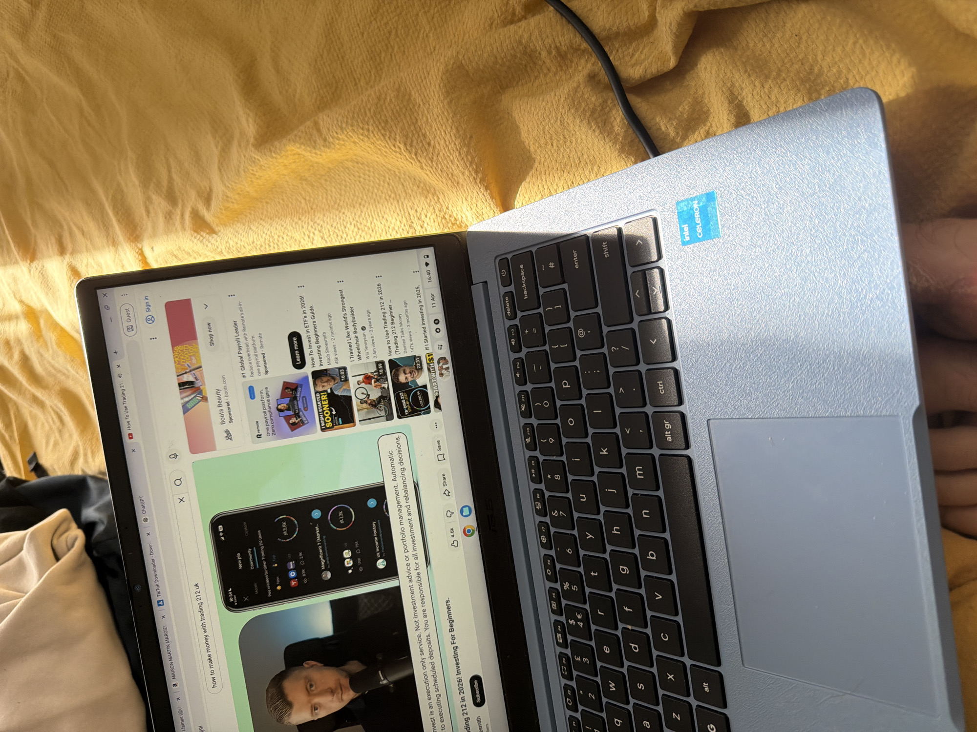 The image shows a light blue laptop on a wrinkled yellow surface, likely a bedspread. The laptop is open, displaying a YouTube page with thumbnails of various videos, including content on investing and finance. The laptop keyboard is visible with the "Intel Celeron" sticker near the top right corner. The screen is lit, and the cursor is visible. A black cord is partially visible in the top right corner of the image, possibly a charging cable. The keyboard is black with gray lettering. Part of a person's leg is visible to the right of the laptop. The image does not depict any location within the city of Pentre, United Kingdom.