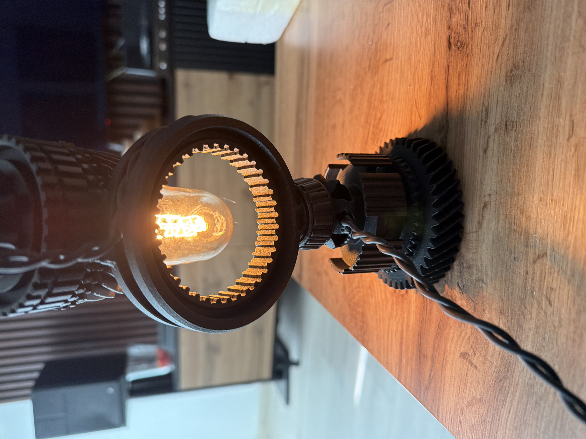 The image captures a close-up view of a unique, industrial-style lamp, oriented vertically. The lamp is constructed from several interlocking gear-like components, finished in a dark, matte color, possibly 3D-printed due to visible textures. At its center, an Edison-style filament light bulb shines brightly, emitting a warm, golden-orange glow. This illumination highlights the intricate teeth along the inner edge of one of the large gear rings that frame the bulb.
The lamp is positioned on a polished wooden surface, likely a desk or shelf, which displays a prominent grain pattern. The warm light from the bulb casts a soft, elongated shadow from the lamp's base onto this wooden surface. A black, twisted electrical cable extends from the lamp's base, trailing across the wooden top towards the lower right of the frame.
The background is blurred and out of focus, showing darker elements on the left, possibly a wall or shelving, and lighter, indistinct shapes towards the upper right. The overall impression is an indoor setting, characterized by a cozy and warm ambiance, typical of evening or nighttime illumination. There are no people or discernible activities beyond the lamp being lit. No text is visible in the image, and no specific location cues for Sofia, Bulgaria, are present within the visual content.