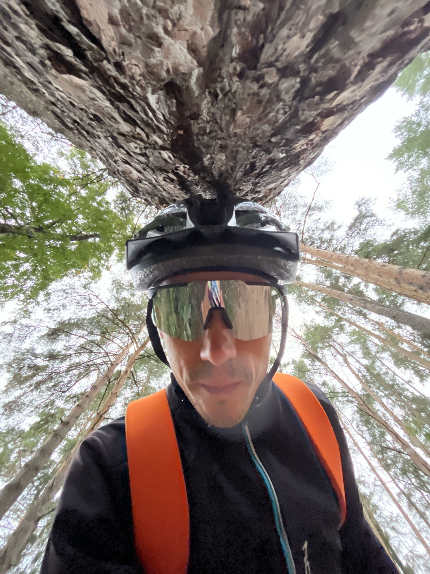 The image, taken from a low-angle perspective, features a person wearing a black cycling helmet and reflective goggles. The person is also wearing a black jacket with orange straps over the shoulders, possibly part of a backpack or hydration pack. In the background, tall, slender trees with green foliage are visible against an overcast sky. A large, textured rock formation overhangs the scene from above. The image appears to be taken in a wooded outdoor environment. There is no specific indication of the city of Chaikovsky, Russia within the image itself.