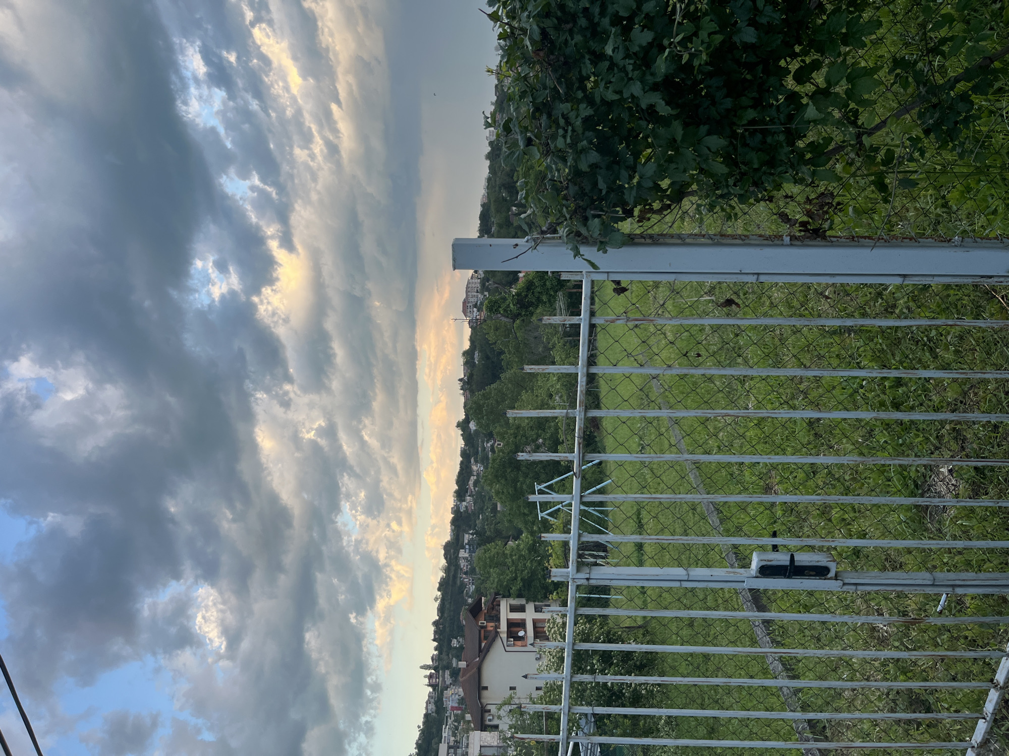 The image shows a view through a metal fence with a green, grassy area behind it, possibly a park or field. In the background, there is a cityscape, including houses and buildings, nestled on a hillside. The sky is cloudy with a mix of dark and light areas. The photo appears to have been taken at an angle. Given the presence of houses and buildings nestled on a hillside, combined with the cloudy sky, the location is likely in the city of Varna, Bulgaria.