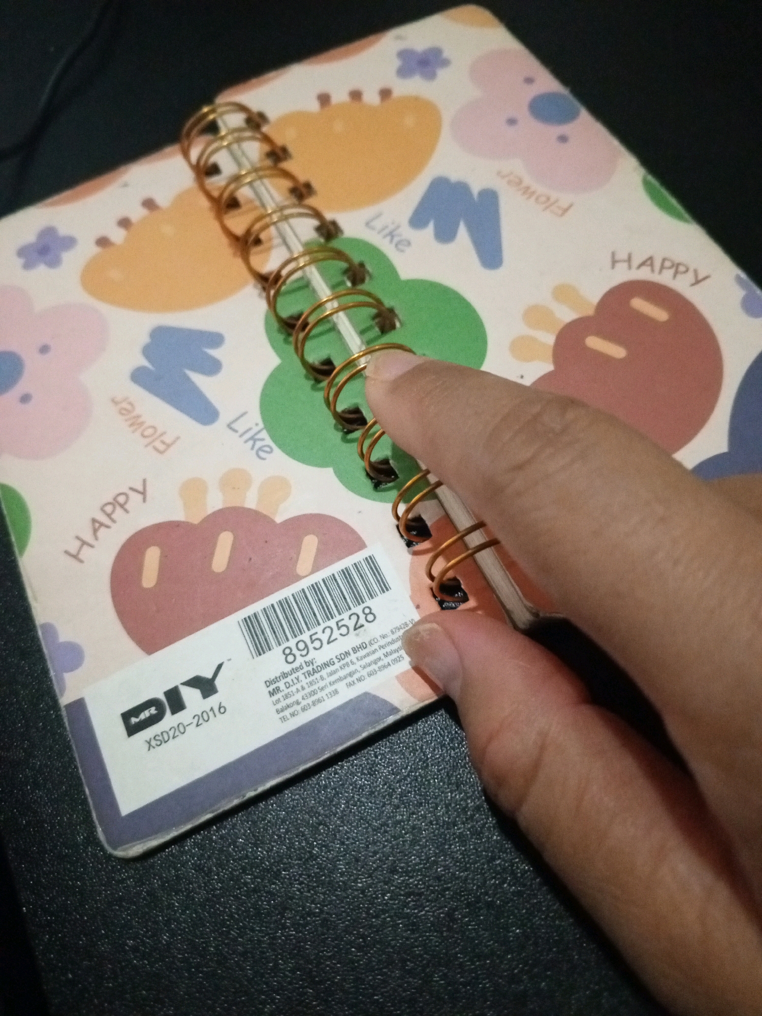 An overhead shot shows a notebook with a playful, colorful design and a spiral binding. The notebook cover features scattered cartoonish shapes, including flowers, abstract blue and pink elements, and two large, stylized brown and pink "blobs" with smaller yellow oval shapes resembling eyes. Some words are printed on the cover in a light pink color: "Like", "Flower" (written vertically and backward), and "HAPPY" (written vertically). A hand is visible on the right side of the image, with a thumb and forefinger gripping the spiral binding of the notebook. The notebook is placed on a dark, textured surface, possibly a table. A white sticker is affixed to the bottom left of the notebook, displaying a barcode, the number "8952528", and distribution information for "MR. D.I.Y. TRADING SDN BHD". Below this, the brand "DIY" is visible in large letters with "MR" to its left, and below that, "XSD20-2016". The background is dark. The image does not provide any information about the city of Denpasar, Indonesia.