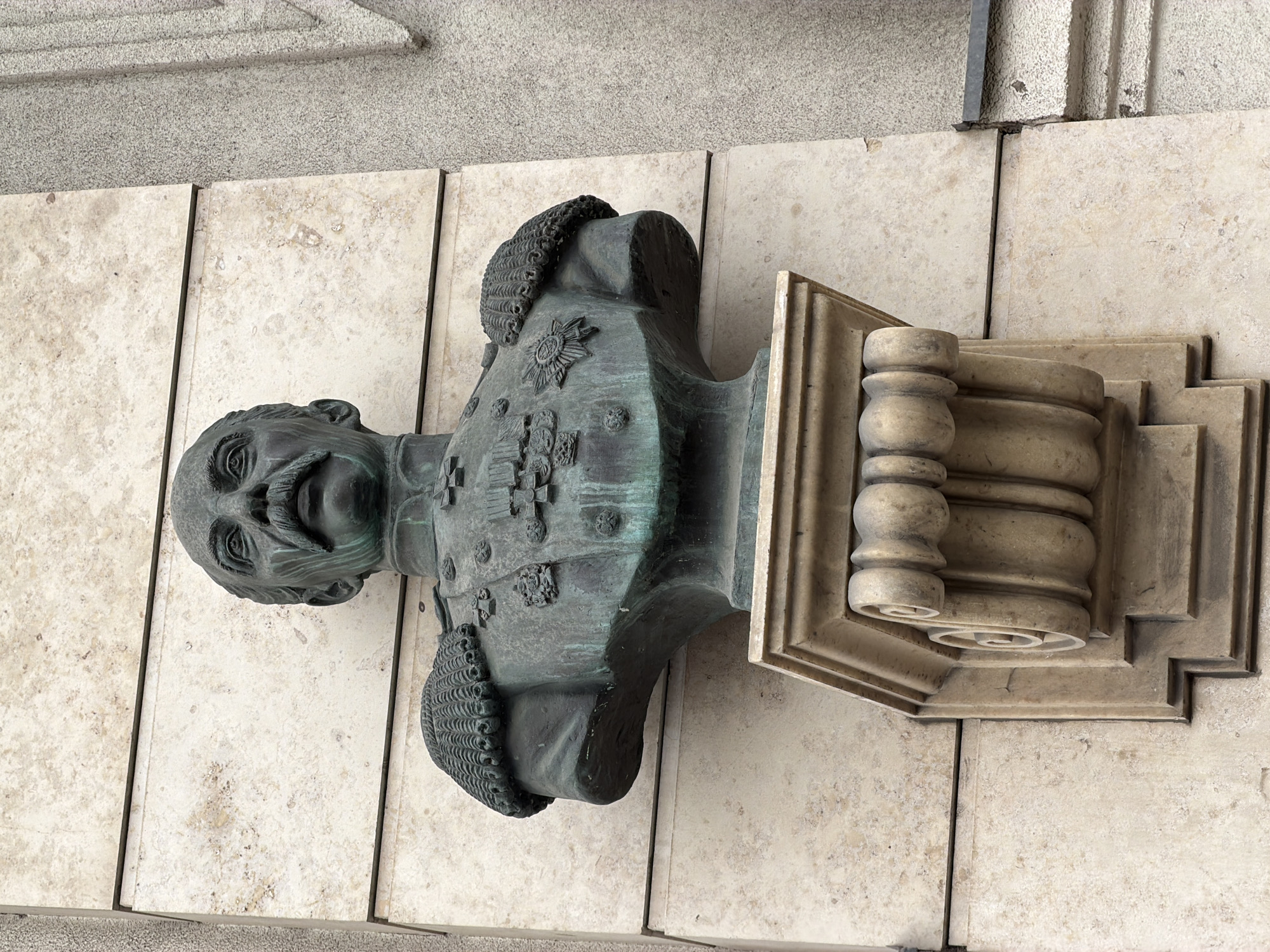 A weathered bronze bust of a military figure with a prominent mustache and ornate uniform is mounted on a decorative stone support attached to a wall composed of vertical marble panels. The bust is placed on a wall in Saint Petersburg, Russia.