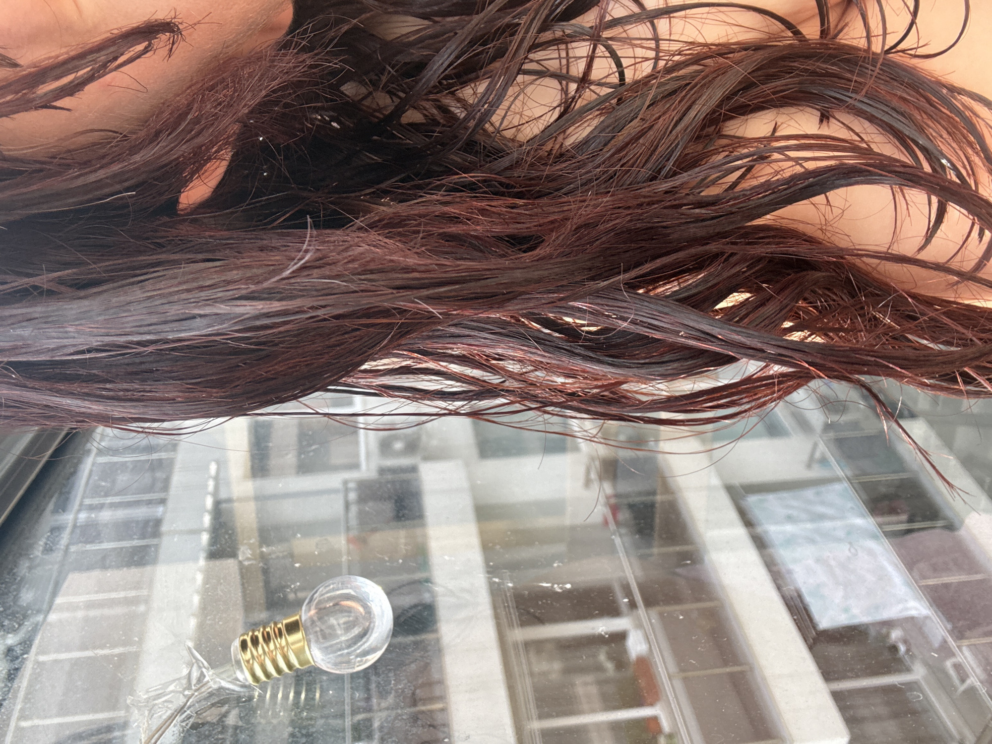 A close-up shot shows a person's wet, dark red hair in the foreground, obscuring much of their face and body. The hair is parted on the right and falls loosely over their shoulder. In the background, through a window, is a view of an apartment building. The window is dirty and has a string of decorative lights hanging on the left side. The lights are shaped like small lightbulbs with gold bases. The apartment building appears to be in an urban setting, with balconies and windows visible. The lighting suggests it is daytime.