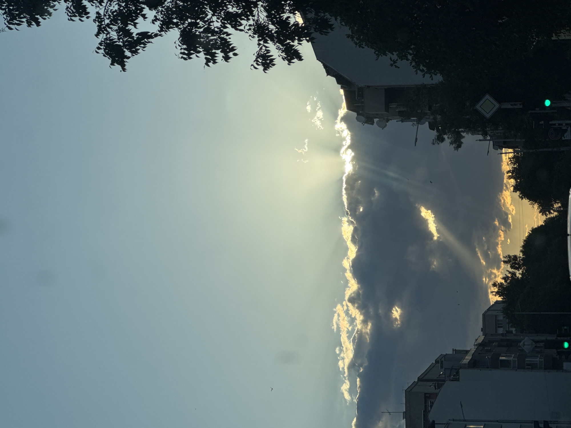 The image captures a street scene in Plovdiv, Bulgaria, likely during the late afternoon or early evening, as indicated by the dramatic lighting. The sky is a soft gradient of blue, transitioning to a yellowish hue near the horizon, partially obscured by a line of dark, heavy clouds. Brilliant sunbeams pierce through the clouds, creating a crepuscular effect.
In the foreground, on the left side, a modern residential building with multiple stories and balconies is visible, its facade illuminated by the diffused sunlight. To the right, a taller, older building with a rougher texture and numerous satellite dishes is silhouetted against the bright sky. Trees, some with dense foliage and others appearing as dark silhouettes, frame the scene on the right and in the distance.
Along the street, a traffic light displays a green signal. A road sign, a white diamond with a yellow border, is also present. The overall impression is one of urban life bathed in the atmospheric glow of a setting sun, with the interplay of light and shadow emphasizing the architectural elements of the city. There are no visible people or animals in the frame.