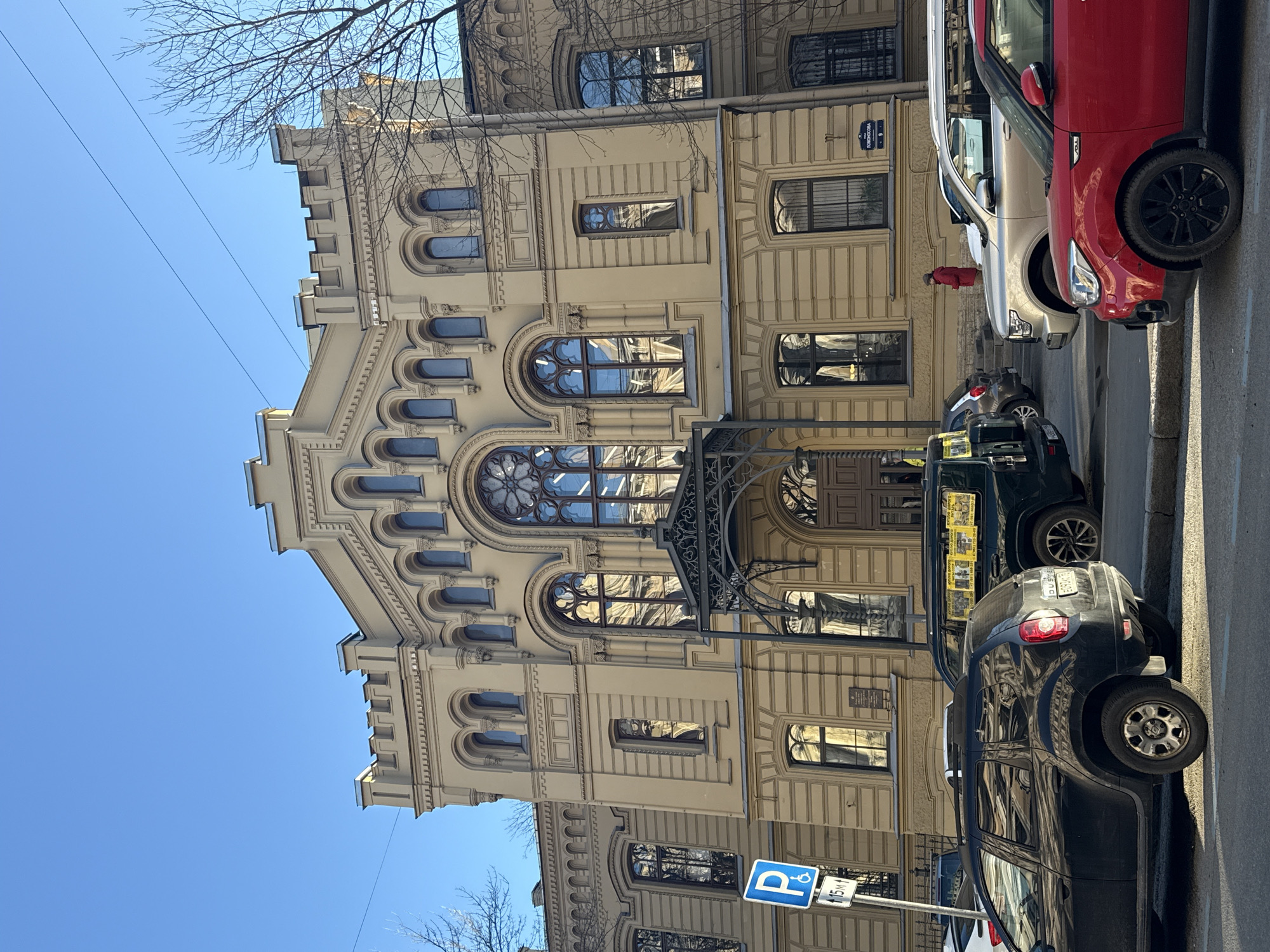 The image captures a sunlit street scene in Saint Petersburg, Russia, featuring a grand, ornate building under a clear blue sky. The multi-story structure dominates the frame, showcasing light beige or cream-colored facades with intricate architectural details. Its design includes numerous arched windows, some featuring decorative tracery reminiscent of rose windows, and a prominent gable with what appear to be crenellated or stepped battlements along the roofline. A dark, ornate metal canopy extends from the building over a ground-floor entrance.
Parked along the street in front of and to the right of the building are several cars. A striking red Kia Soul is visible on the right, followed by a silver SUV. Further down the street, a black SUV is parked, with part of another vehicle behind it. The reflections of the building and sky are visible in the cars' windows. A small red fire hydrant stands on the sidewalk.
A blue square parking sign, displaying a white "P" and a wheelchair symbol, along with "15M," is clearly visible in the bottom right corner, indicating a parking area. Bare tree branches are visible against the bright sky at the top and bottom of the frame, suggesting a late winter or early spring day. Thin power lines cross the upper left sky.
On a building adjacent to the main structure, a street sign with Cyrillic text reads "ПОРОХОВАЯ" (Porokhovaya) and the number "9," confirming the street name. A faint reflection of a person in a red jacket can be seen in the silver SUV's window. The bright light and clear sky suggest it is daytime, likely morning or afternoon. The overall atmosphere is calm and depicts a typical urban environment.