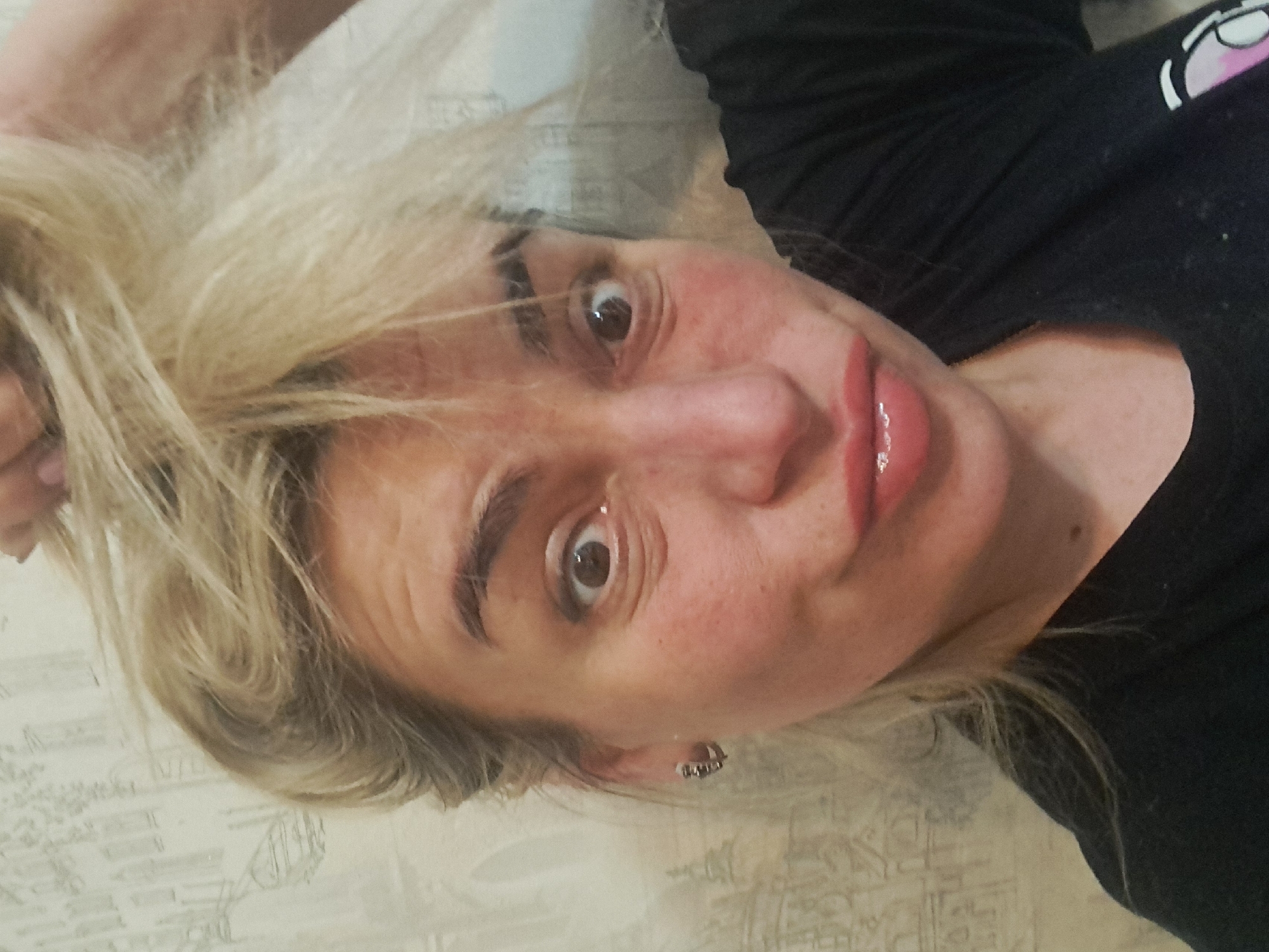 A close-up portrait photograph depicts an adult female positioned indoors in Elektrostal, Russia. The subject has blonde hair, which is styled in a voluminous, disheveled manner, with an unseen hand at the top right of the frame appearing to hold or interact with a portion of it. She faces the camera directly, displaying wide, open brown eyes and puckered lips, conveying a surprised or exaggerated expression. Her facial skin exhibits a fair complexion with reddish undertones, and she wears defined eyebrows, subtle eye makeup, and glossy pink lipstick. A silver-colored hoop earring is visible in her left ear. She is wearing a dark, short-sleeved top, likely a t-shirt. The background consists of an indoor wall covered with light-colored wallpaper featuring a repeating pattern of thin, dark, stylized linear designs resembling architectural outlines or abstract figures.