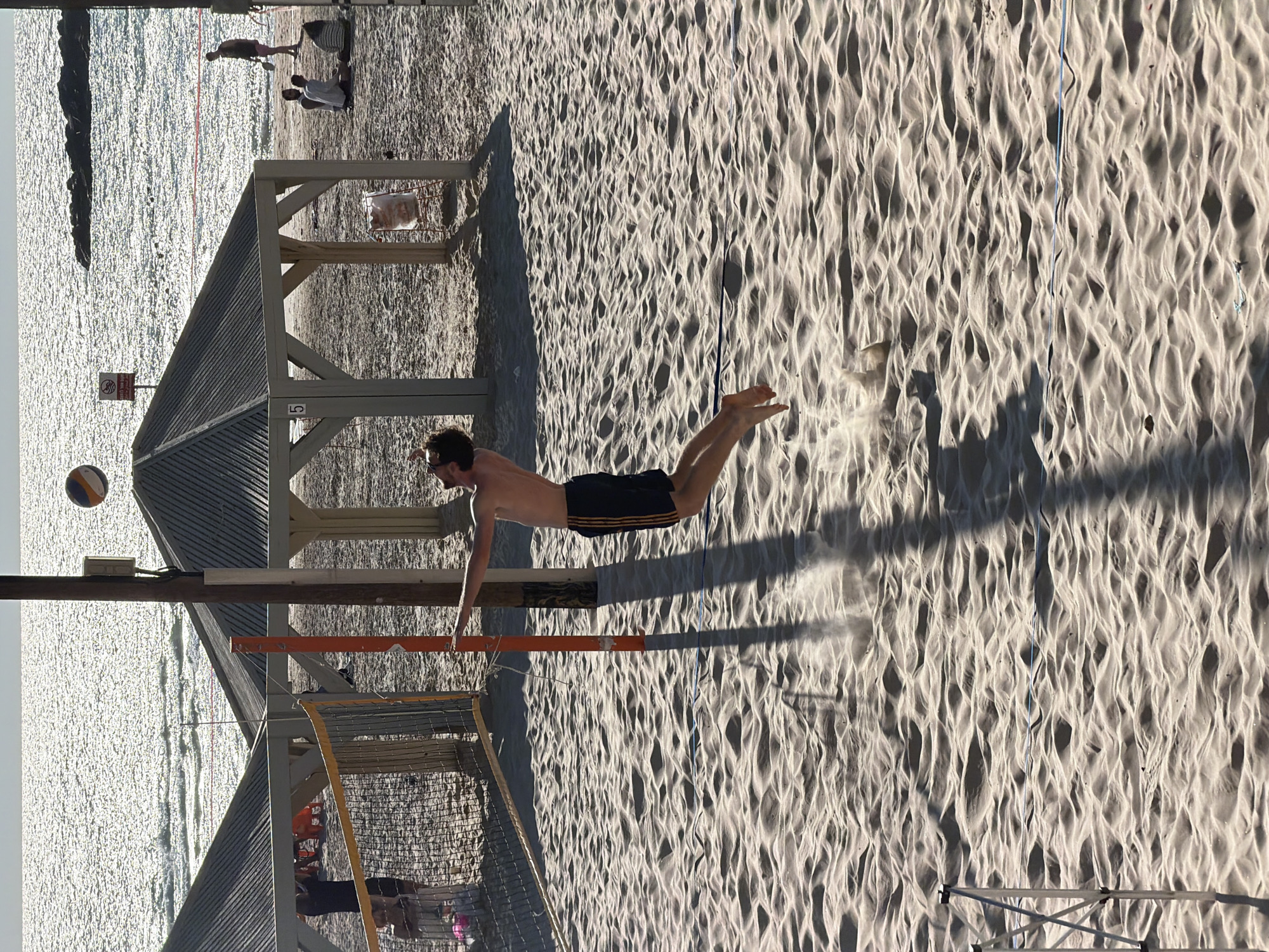 This image captures a vibrant beach volleyball scene, likely in Tel Aviv, Israel, illuminated by bright sunlight that casts long shadows, suggesting either late afternoon or early morning.
In the foreground, a shirtless man wearing dark swim trunks with orange stripes is frozen mid-air, horizontally positioned as if diving or making a play. Sand dust billows around his hands, indicating recent contact with the ground. A white and blue/orange volleyball is visible in the upper left quadrant, implying it is part of the ongoing game. The beach sand features distinct, wave-like ripple patterns, and a thin blue line delineates part of the court. Long, dark shadows from the volleyball net's posts stretch across the textured sand.
A wooden beach volleyball net structure, with orange top bars, stands in the midground. Behind it, a wooden shelter with a sloped roof is visible, marked with the number "5" on its side. Adjacent to the shelter, a white sign with a red circular "no" symbol is partially visible, likely indicating a restriction such as "no swimming" or "no entry."
The background reveals a sparkling expanse of water, presumably the Mediterranean Sea, reflecting the bright sunlight. Several other individuals are scattered along the water's edge; some are standing, others are seated on the sand, observing or relaxing. The overall atmosphere of the image is energetic and bright, depicting a sunny day at the beach.