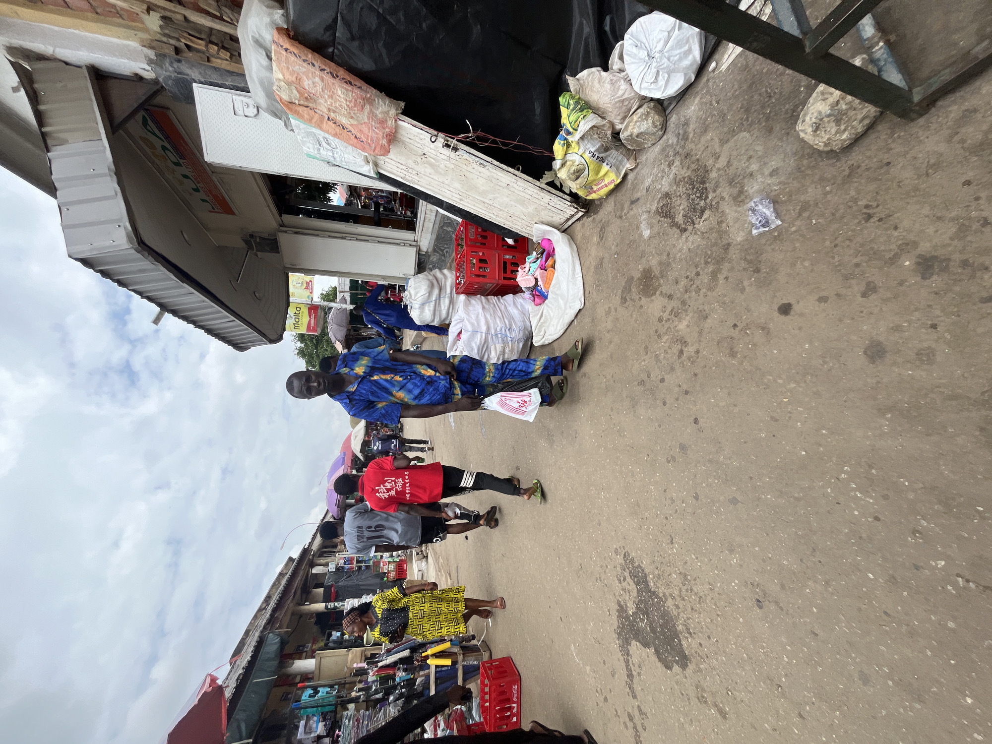The image depicts a bustling outdoor market scene in Abuja, Nigeria. Several people are present, with a prominent focus on a man in a vibrant blue and yellow patterned shirt and pants, carrying a white plastic bag. Other individuals are seen walking and browsing. The setting is a street lined with small shops and stalls, with various goods displayed, including bags of what appear to be rice, red plastic crates, and bundles of clothing. A truck bed is visible in the background, filled with items. The ground is paved, showing signs of wear and some spilled liquids. The sky above is cloudy, suggesting it might be an overcast day. The overall atmosphere appears lively and active, typical of a market environment. Some text is visible on a shop sign, though it is partially obscured and difficult to read. Another sign in yellow with "malta" is visible. The ground also has some unidentifiable text written in white.
