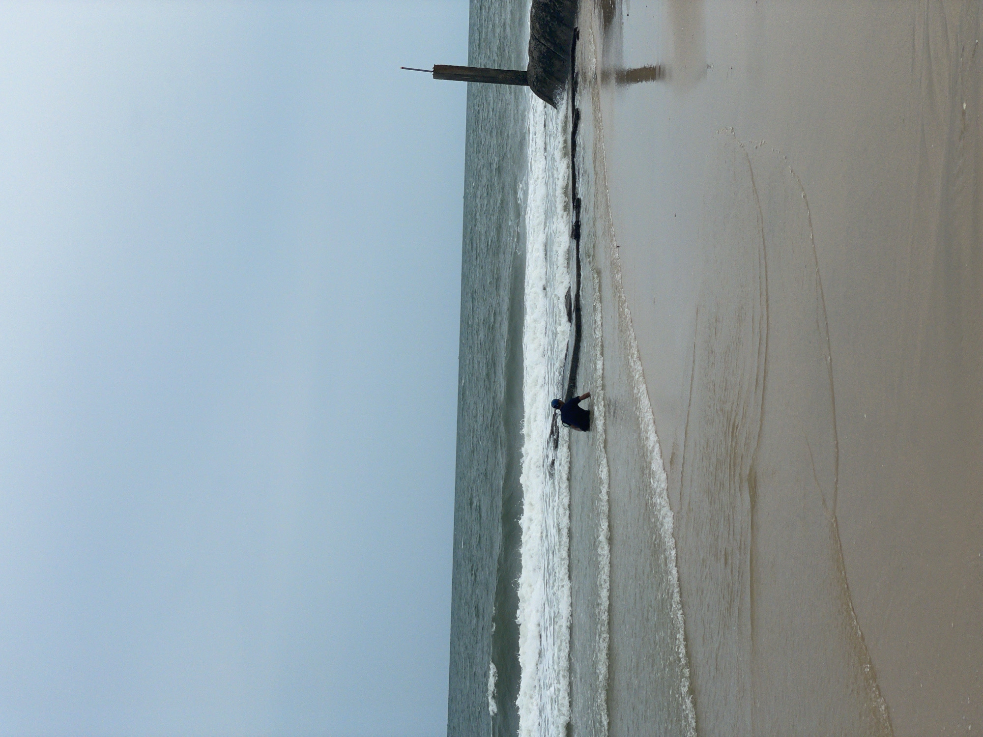 A beach scene in Phu Thuy Ward, Vietnam, features a concrete structure at the water's edge, with a person standing near the waves on the sand. The ocean stretches towards the horizon, and the sky is a pale blue.