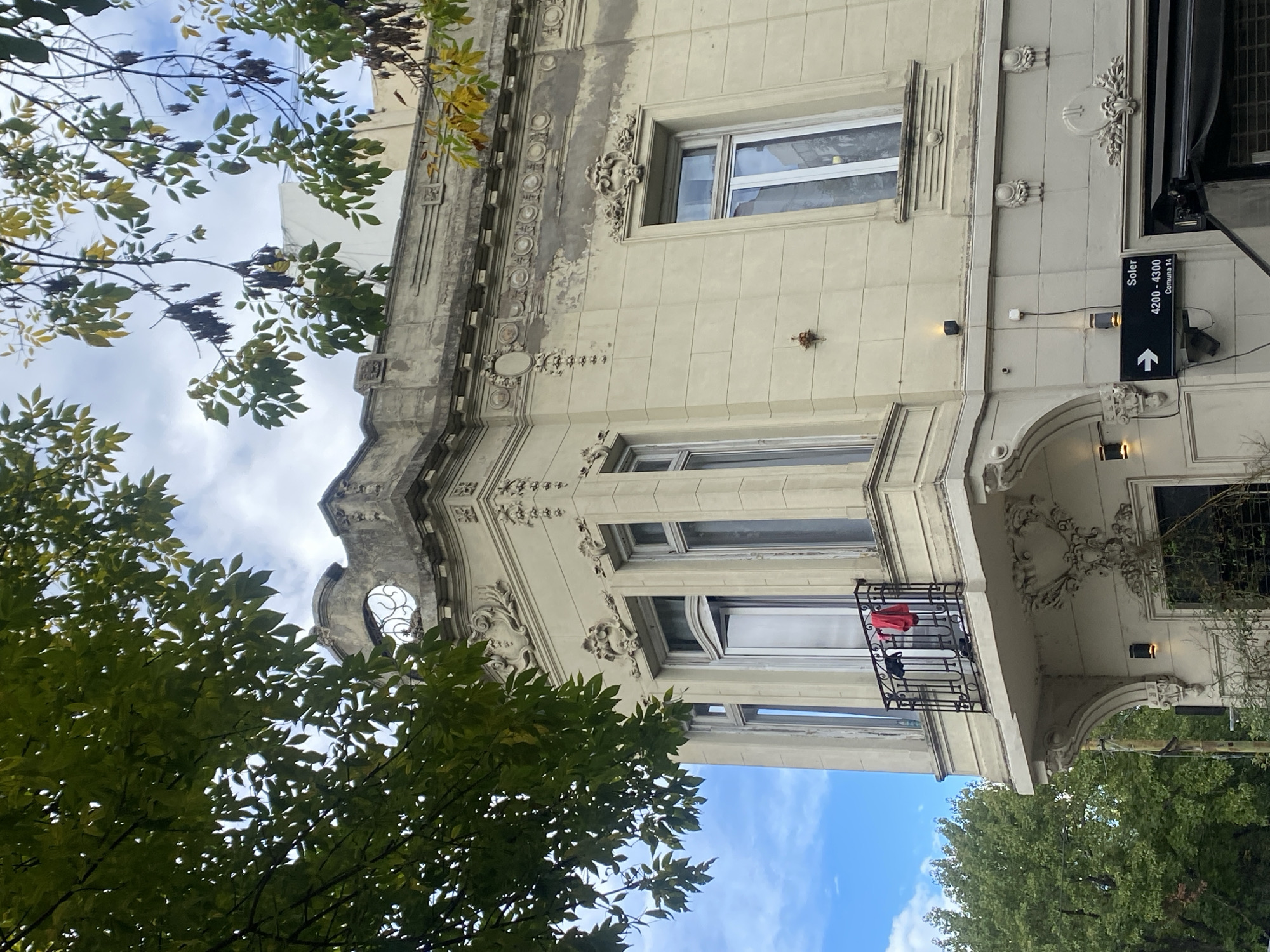 Here's a description of the image:
The image depicts an ornate building in Buenos Aires, Argentina. The building's facade is cream-colored with decorative moldings and several windows. A small balcony with wrought iron railing juts out from the second story, holding clothing. A street sign on the building reads "Soler 4200-4300 Comuna 14" indicating a specific location within the city. Trees with green and yellowing leaves frame the building on both sides, partially obscuring it from view, and the sky is visible between the branches. The image is shot from a low angle, looking upward.