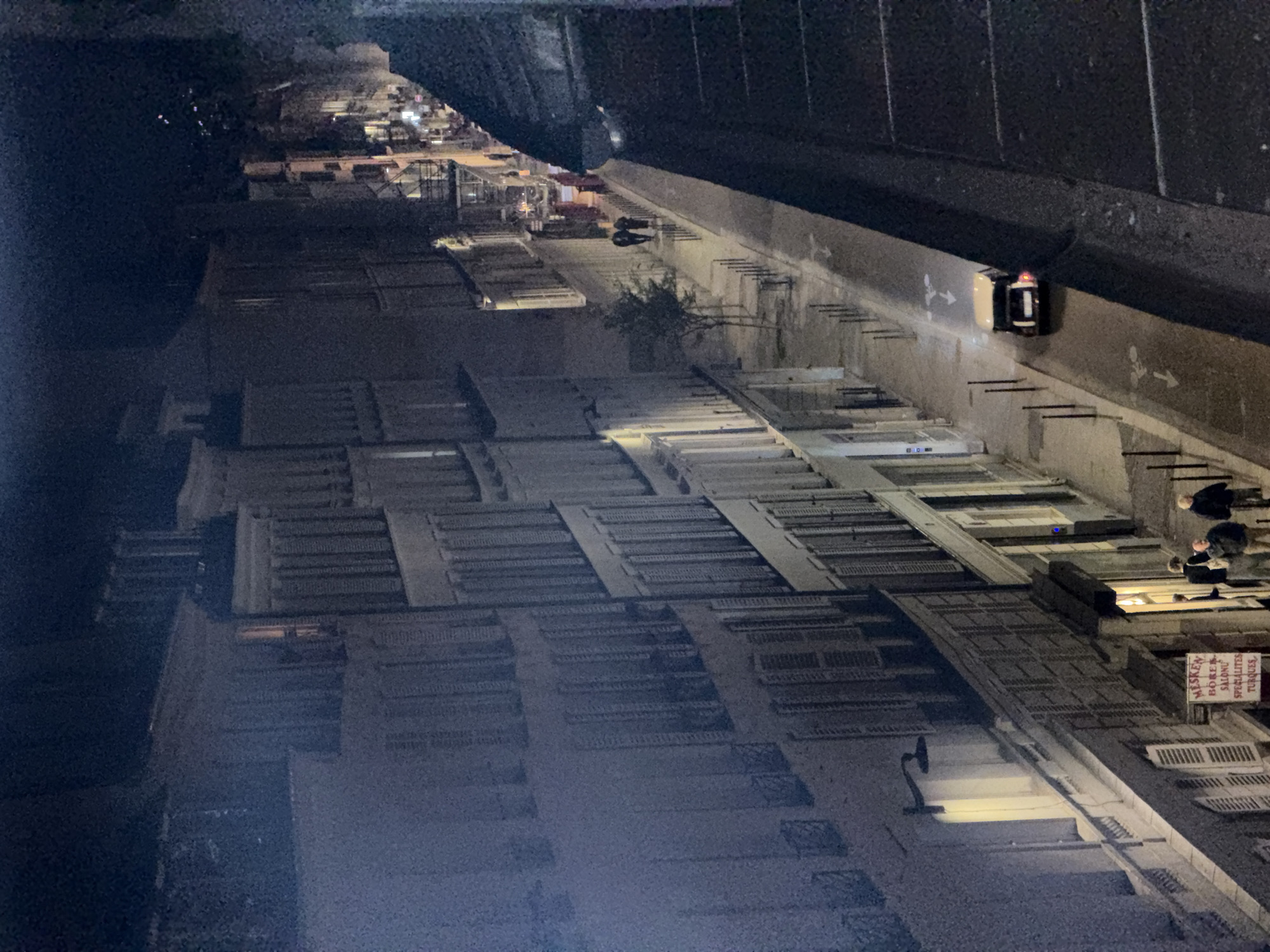 Here's a description of the image:
The image appears to be taken from a high vantage point, looking down at a street in Paris, France. The angle is quite steep, with the buildings stretching into the distance.
**Buildings:** Predominantly residential buildings line the street. They have a consistent architectural style, with numerous windows, many of which are shuttered. The buildings are generally light in color, perhaps off-white or gray.
**Street:** The street is illuminated by streetlights. Pedestrians are visible walking along the sidewalk. Some people are standing near a sign, suggesting a point of interest or a storefront. A single car with its rear lights on is parked alongside the side of the road. The shadows indicate that there may be other objects or structures not fully visible in the photo.
**Overhead Structure:** A dark, heavy structure (likely a bridge or elevated road) appears at the top of the image, partially obscuring the scene.
**Overall Environment:** The image conveys a sense of a quiet evening in Paris. The street is relatively active, but the overall impression is peaceful and well-lit. The presence of the overhead structure adds a sense of enclosure to the scene. The construction site visible in the distance suggests some activity in the vicinity.