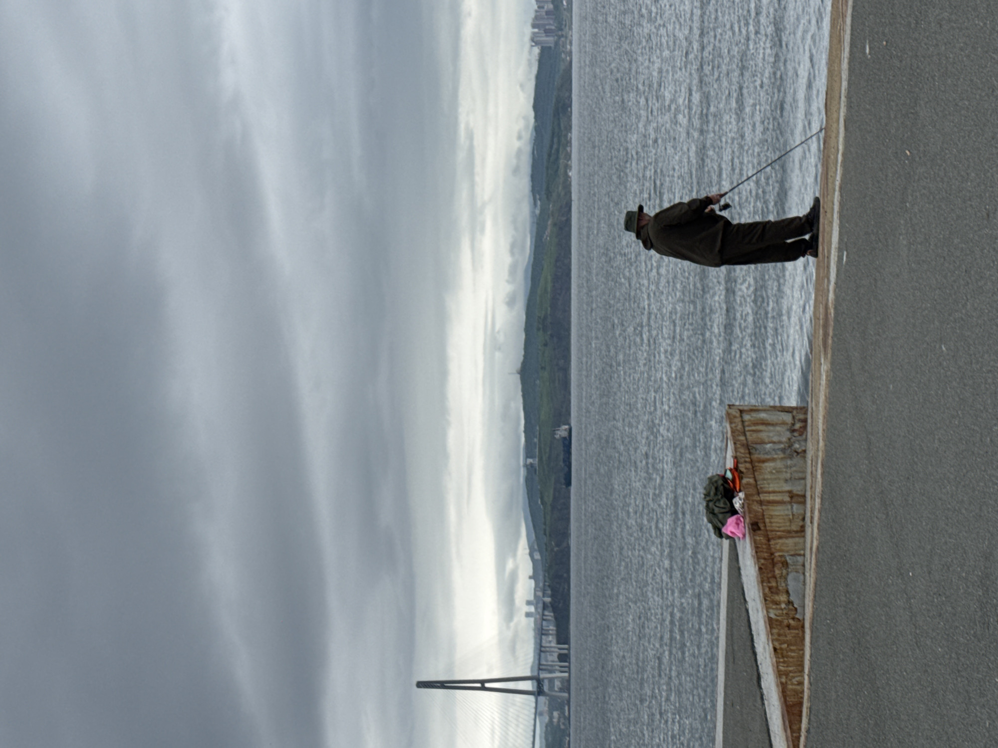 An overcast, grey sky hangs over a calm, grey sea in this full vertical shot. The foreground features an asphalt surface leading to a concrete pier with rusted metal edges. On the pier, a pile of dark clothing and a bright pink object are visible. In the mid-ground, a lone figure, a man wearing a hat and dark clothing, stands facing the water, holding a fishing rod. His posture suggests he is fishing. In the background, across the water, a large cable-stayed bridge with a prominent central pylon dominates the horizon. Beyond the bridge, a green, hilly landscape is dotted with buildings, indicating a city. A cargo ship is visible in the water between the shore and the bridge, and further in the distance, more city structures can be seen along the coastline. The overall impression is one of a quiet, somewhat somber day by the sea in a coastal urban environment, possibly Russky Island, Russia, given the distinctive bridge architecture.